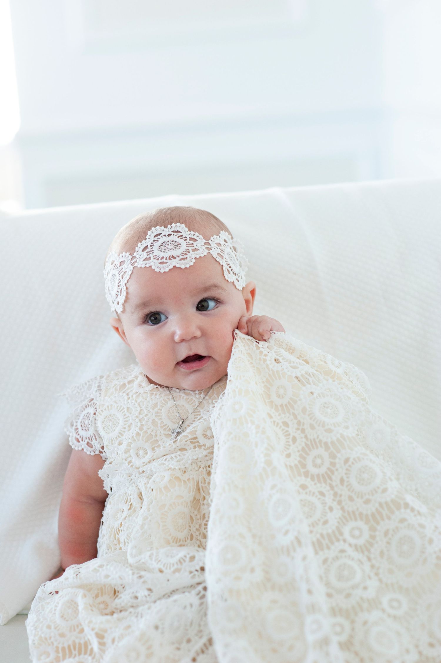 Baby wearing white lace dress and headband lying on white fabric.