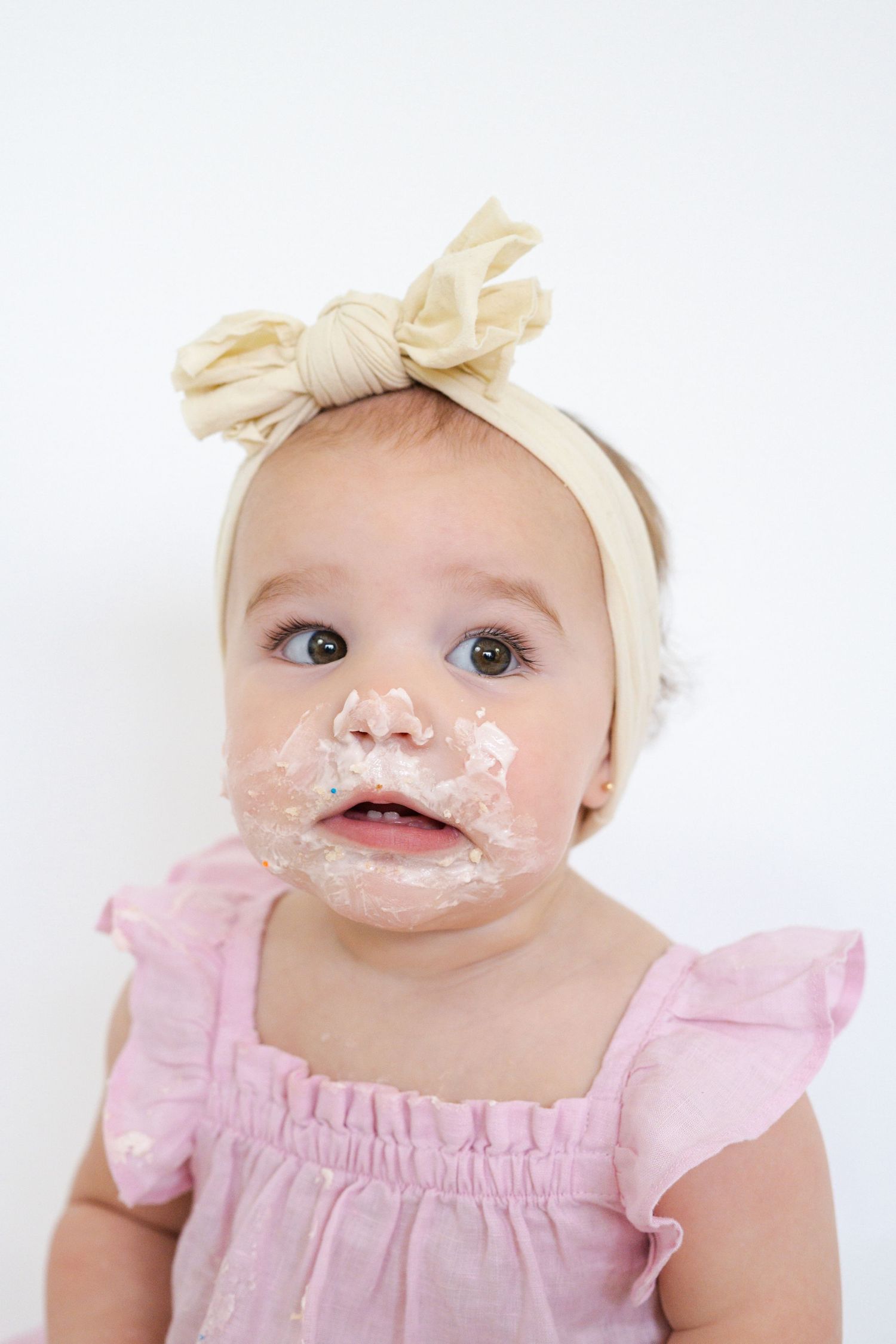 Baby wearing cream bow and pink dress with cake frosting on face during first birthday session.
