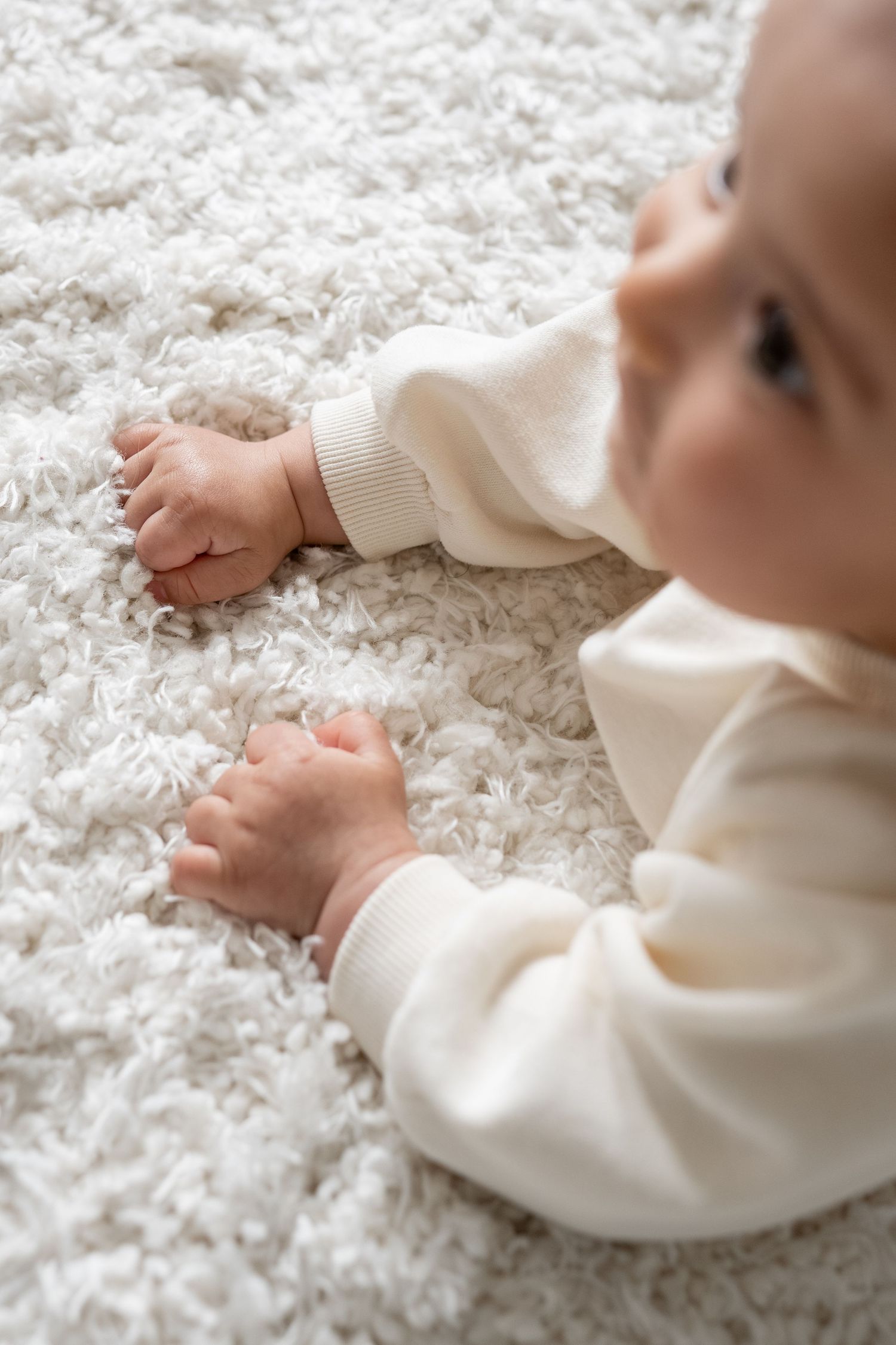 Close-up of baby in cream onesie crawling on soft white shag carpet with tiny bare feet visible.
