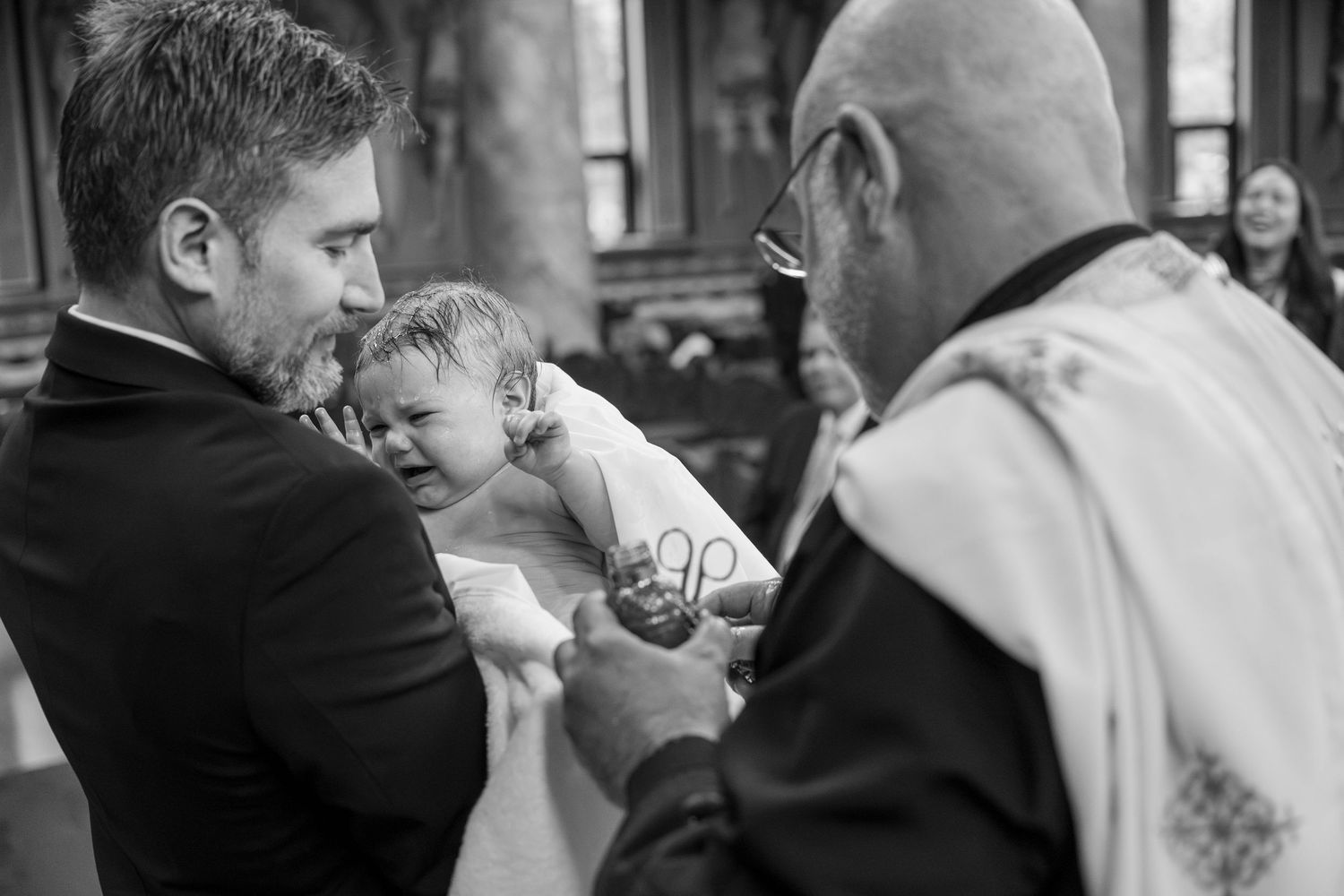 Black and white photo of a crying baby during a baptism ceremony, held by father while priest performs the christening.