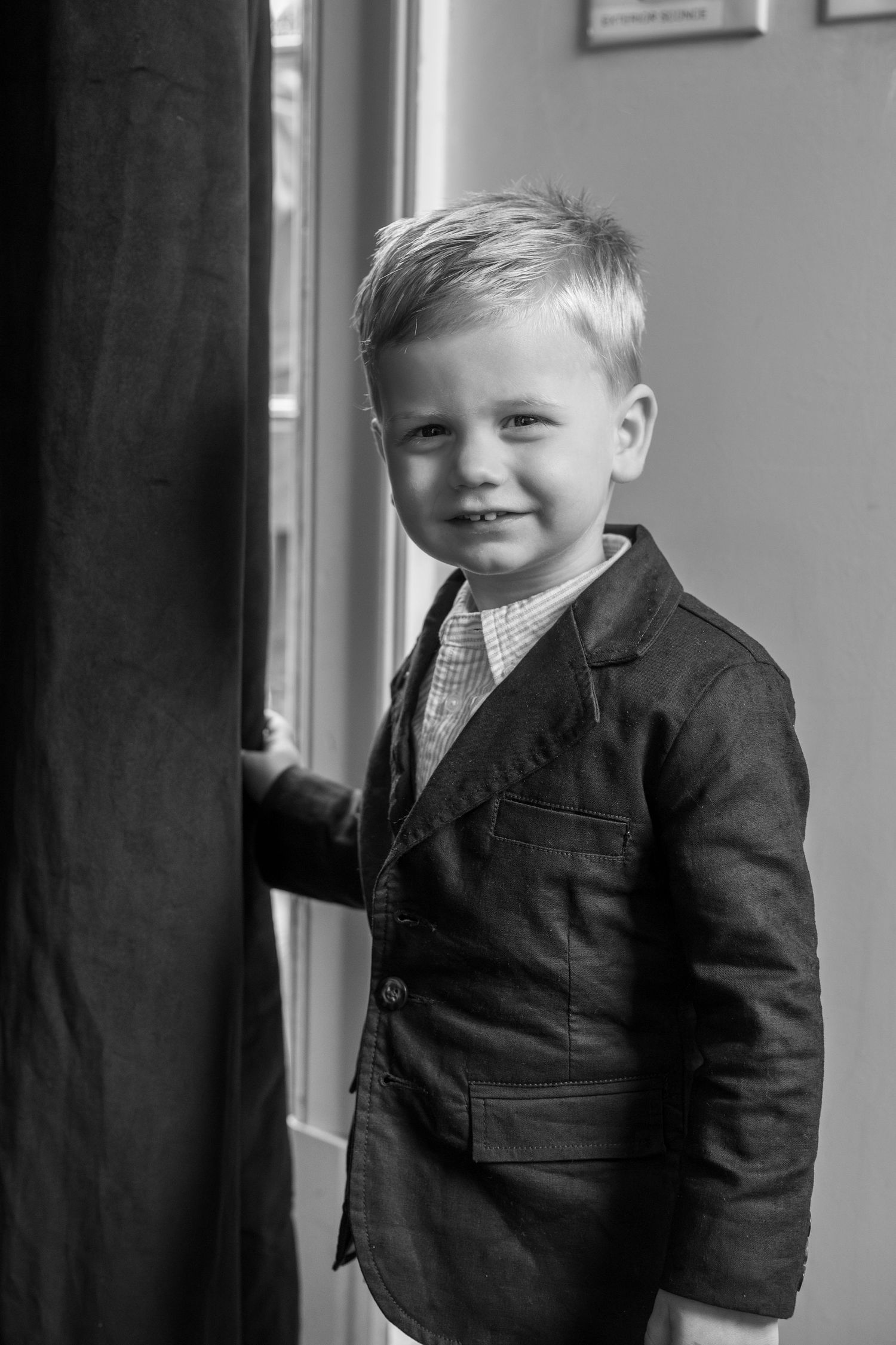 A young boy in a blazer smiles beside a curtain near a window in a classic black and white portrait photo.
