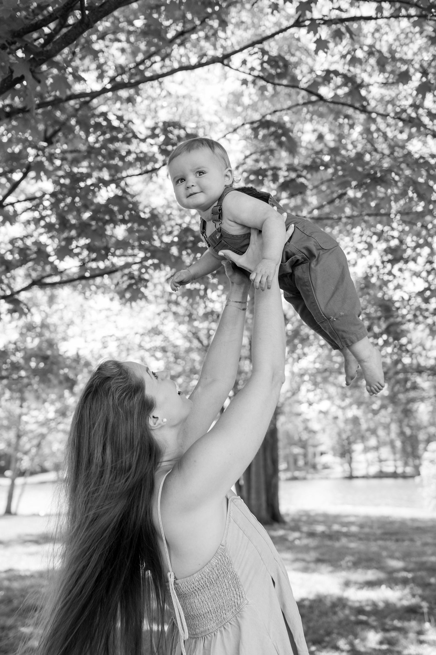 Black and white photo of a mother lifting her smiling baby up in the air outdoors among trees.
