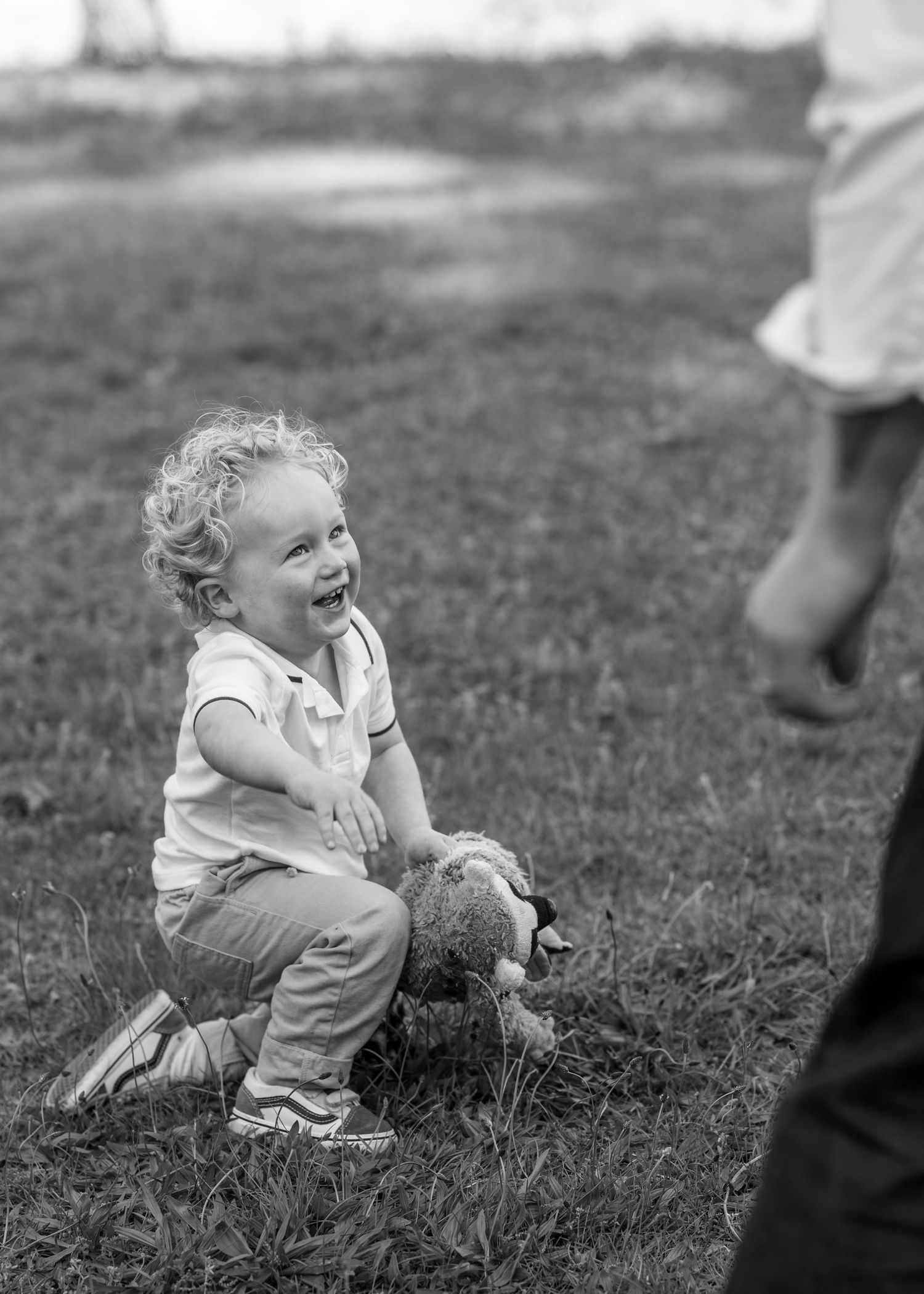 Black and white photo of a laughing toddler crouching on grass holding a stuffed animal toy outdoors.