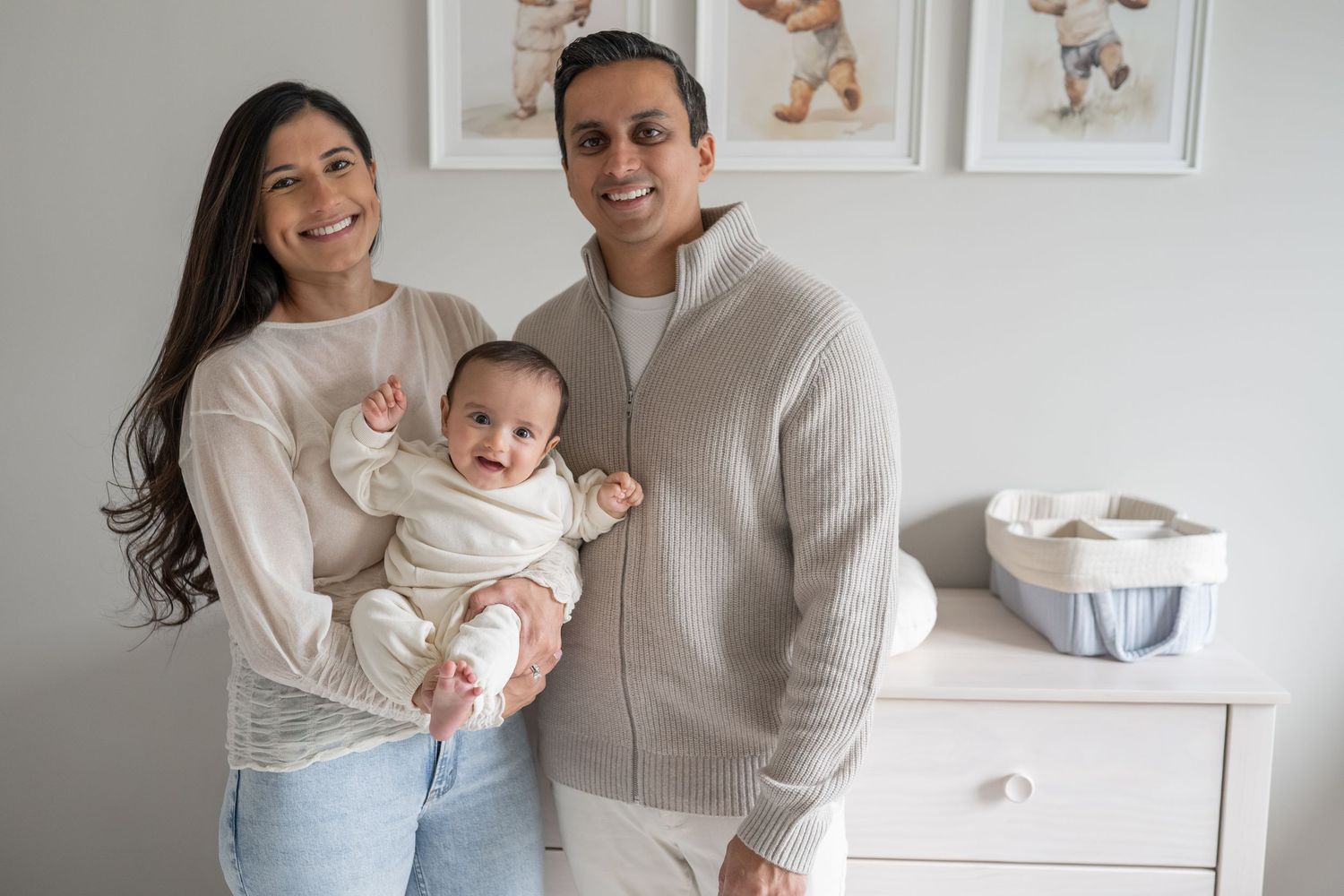 A family in neutral colored sweaters poses together in a nursery with framed artwork and white furniture.