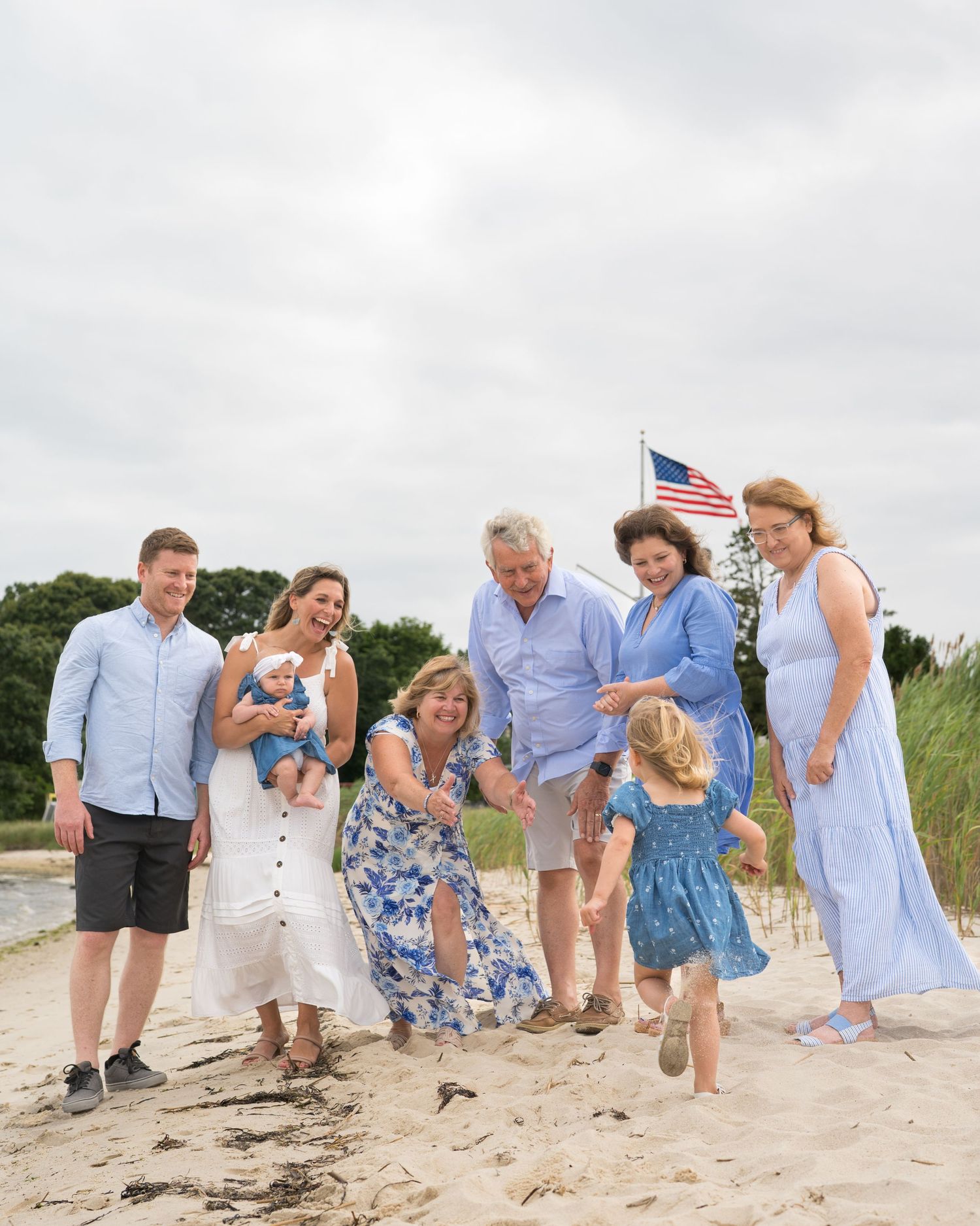 Family group playing and running together on a sandy beach with an American flag in the background.