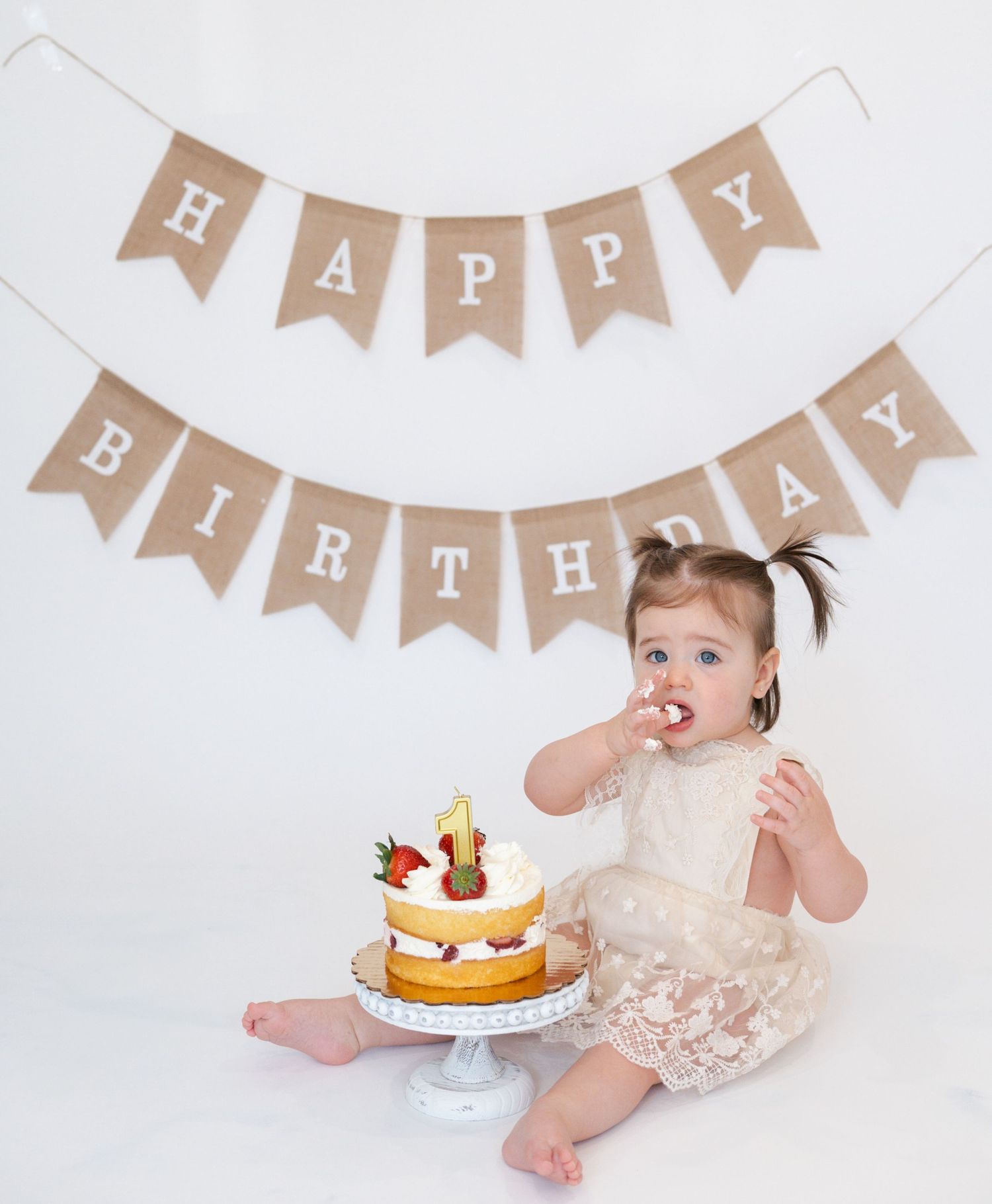 Baby in lace dress reaches for birthday cake with banner decoration.