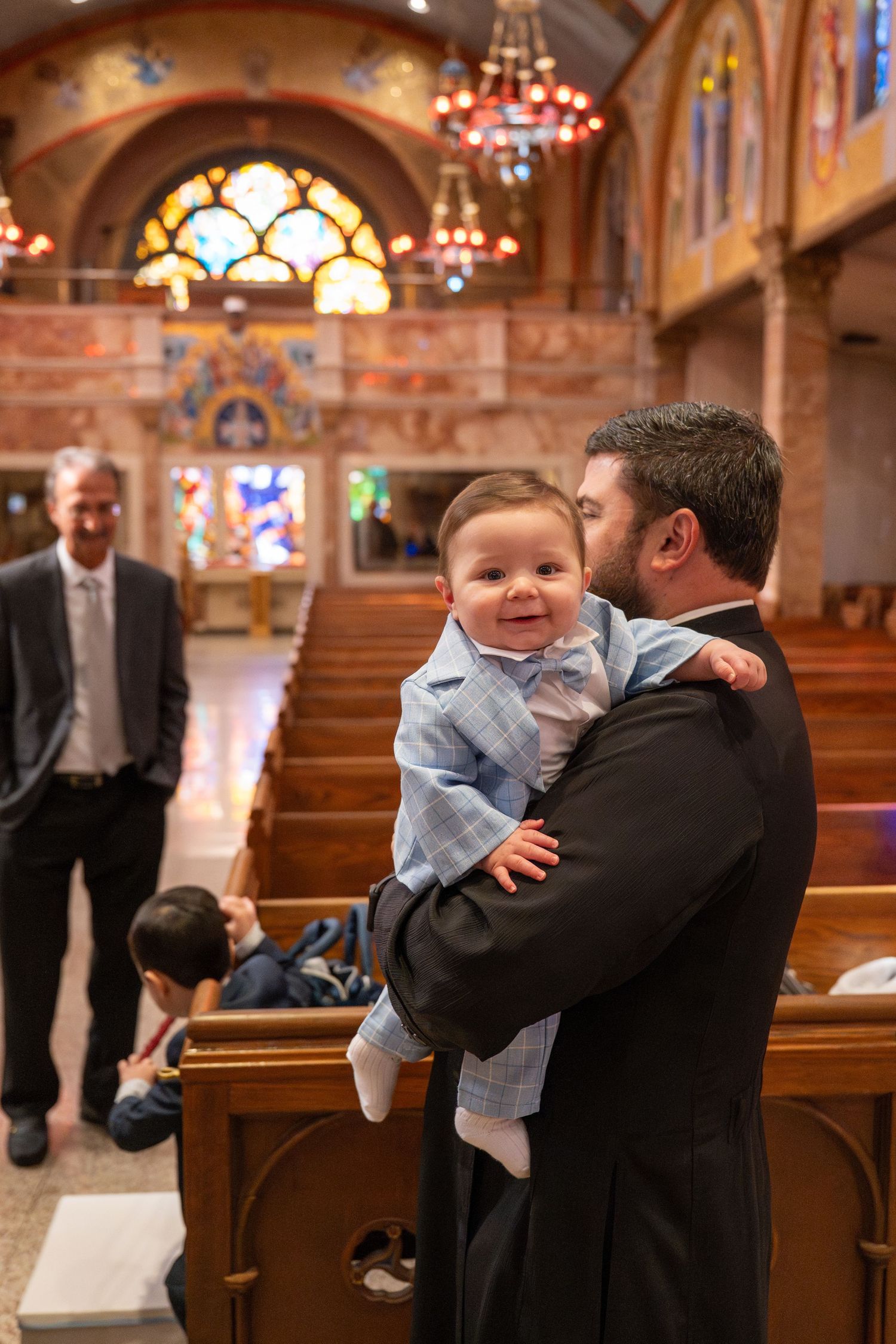 A joyful moment between a baby in blue and an adult in formal wear inside an ornate church with stained glass windows.