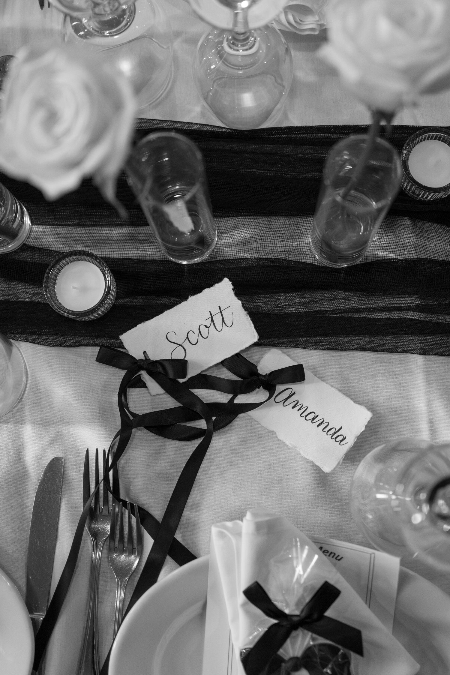 Black and white photo of wine glasses and place cards with ribbons arranged on a table at an elegant event.