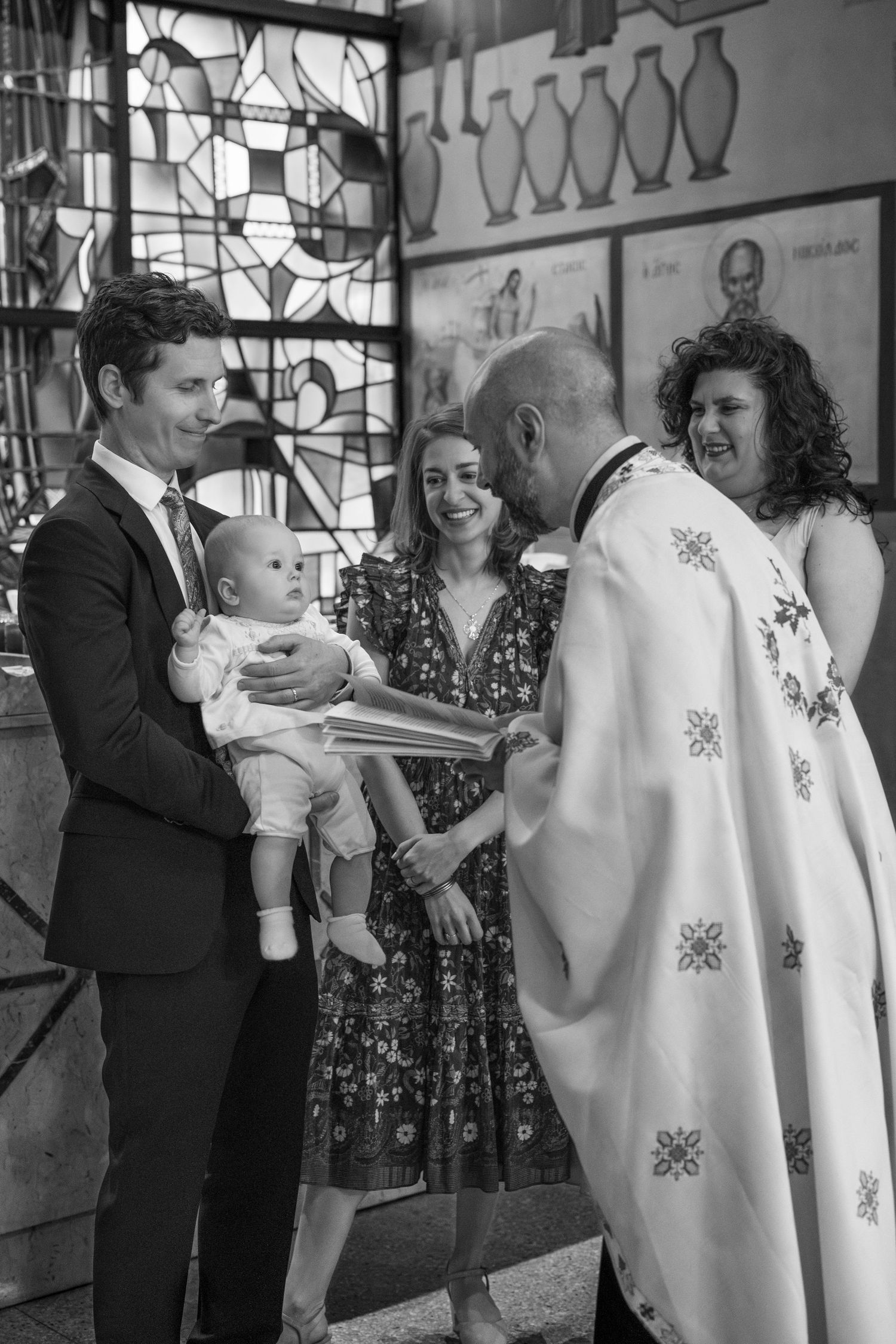 A black and white photograph of a baptism ceremony in a church with a priest wearing traditional robes.