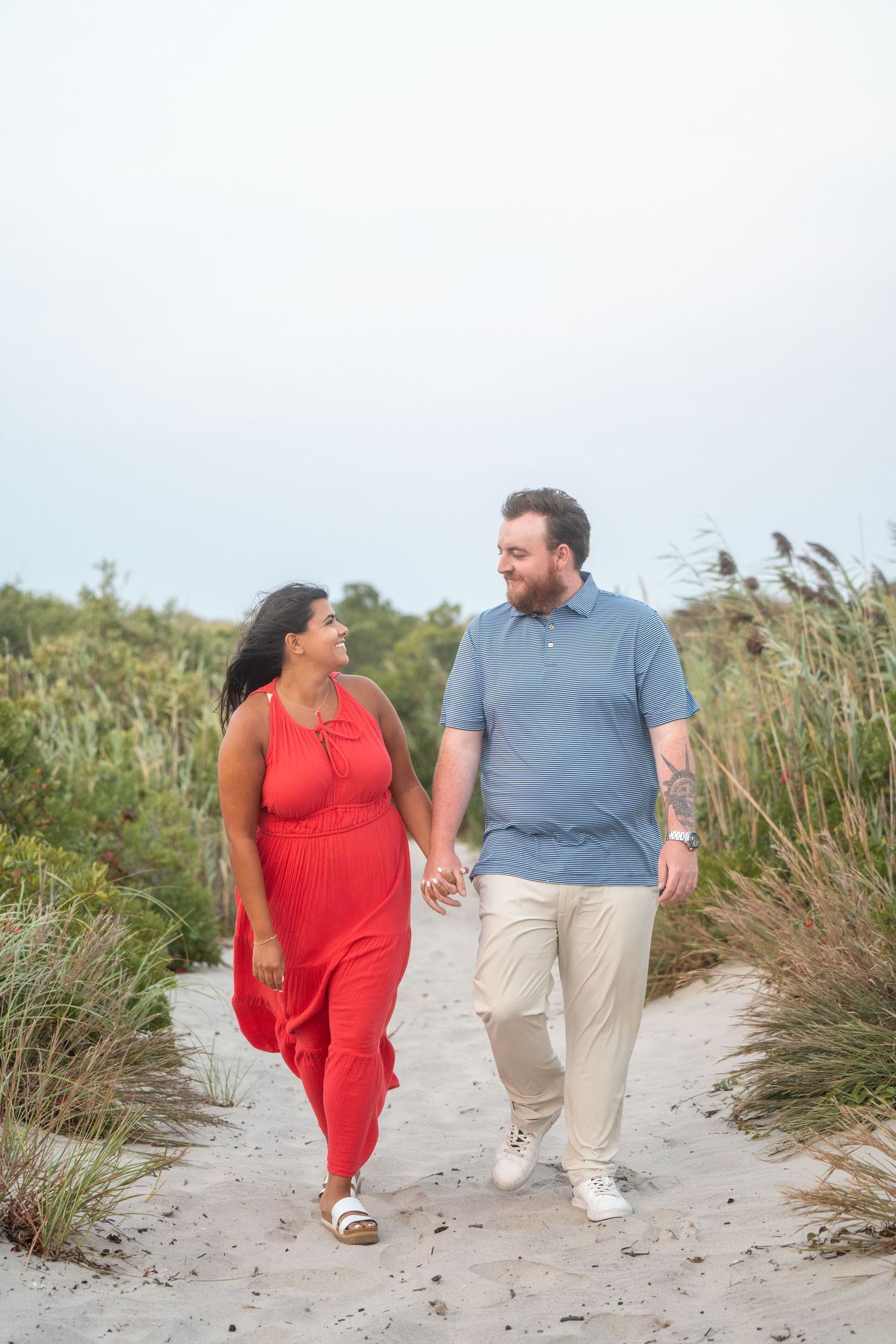 A couple walks hand in hand along a sandy beach path lined with sea grass on a sunny day.