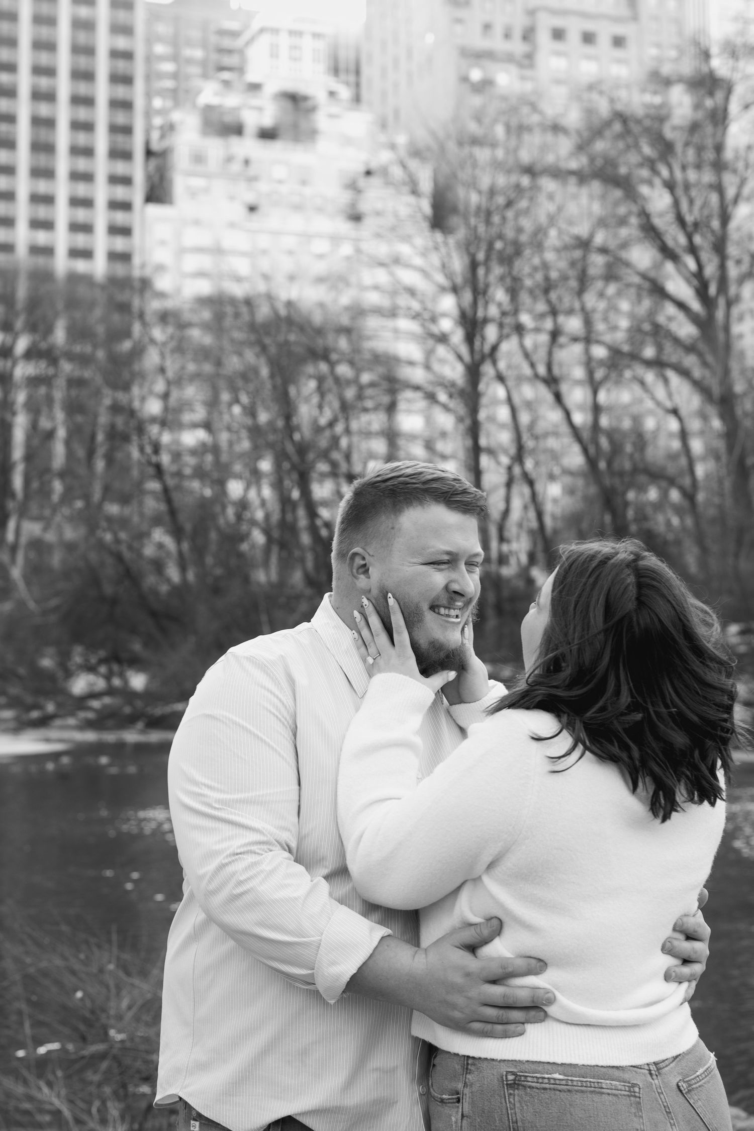 A joyful couple embraces and laughs together in a winter park setting with city buildings behind them.