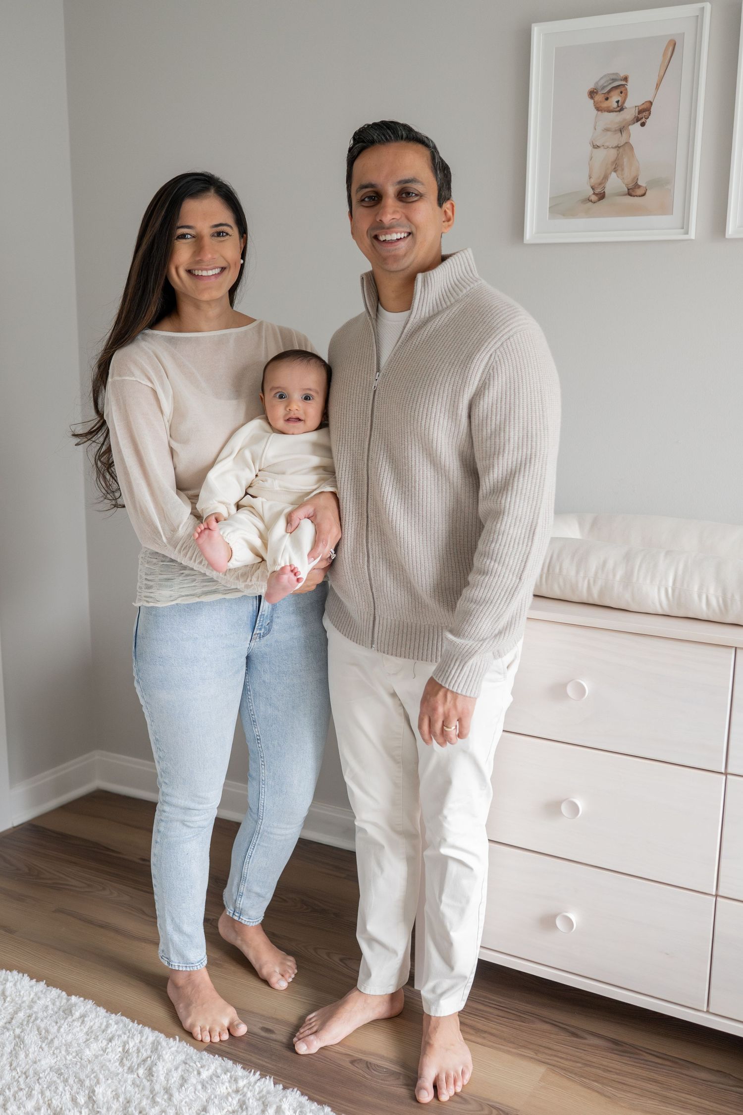 Family in neutral-toned outfits poses together in front of white furniture in a minimalist nursery setting.