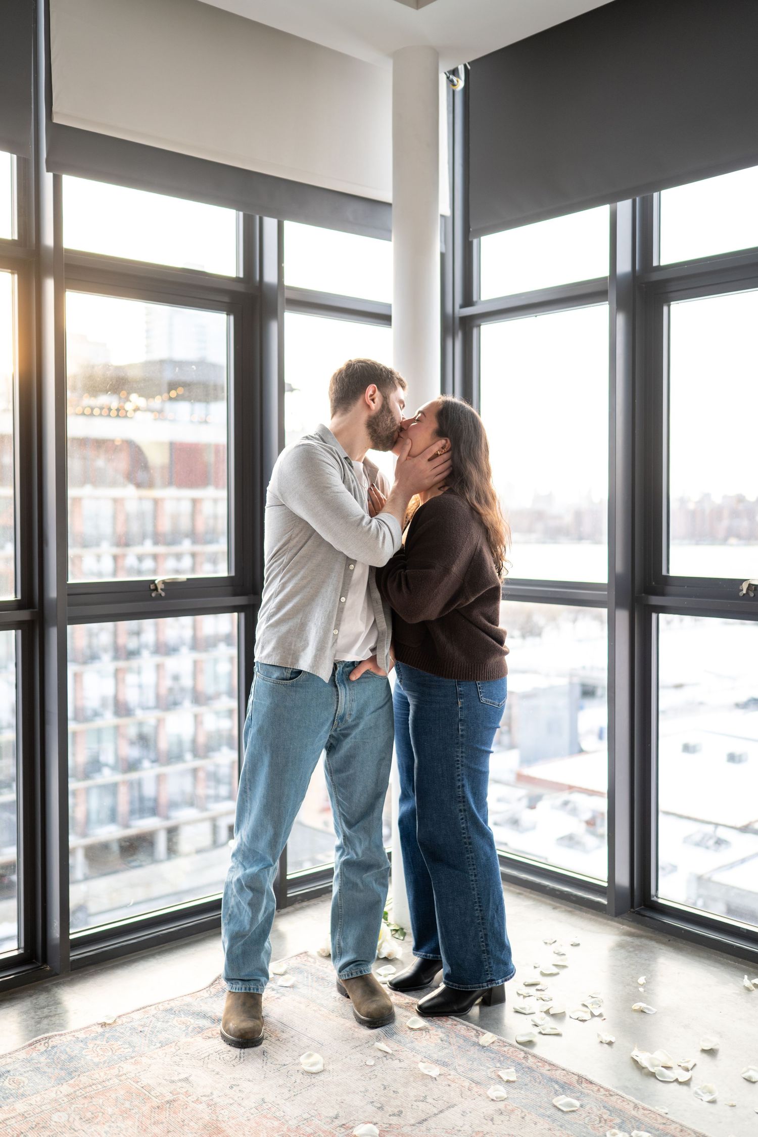 A couple embracing in front of large windows in an empty room with city views.