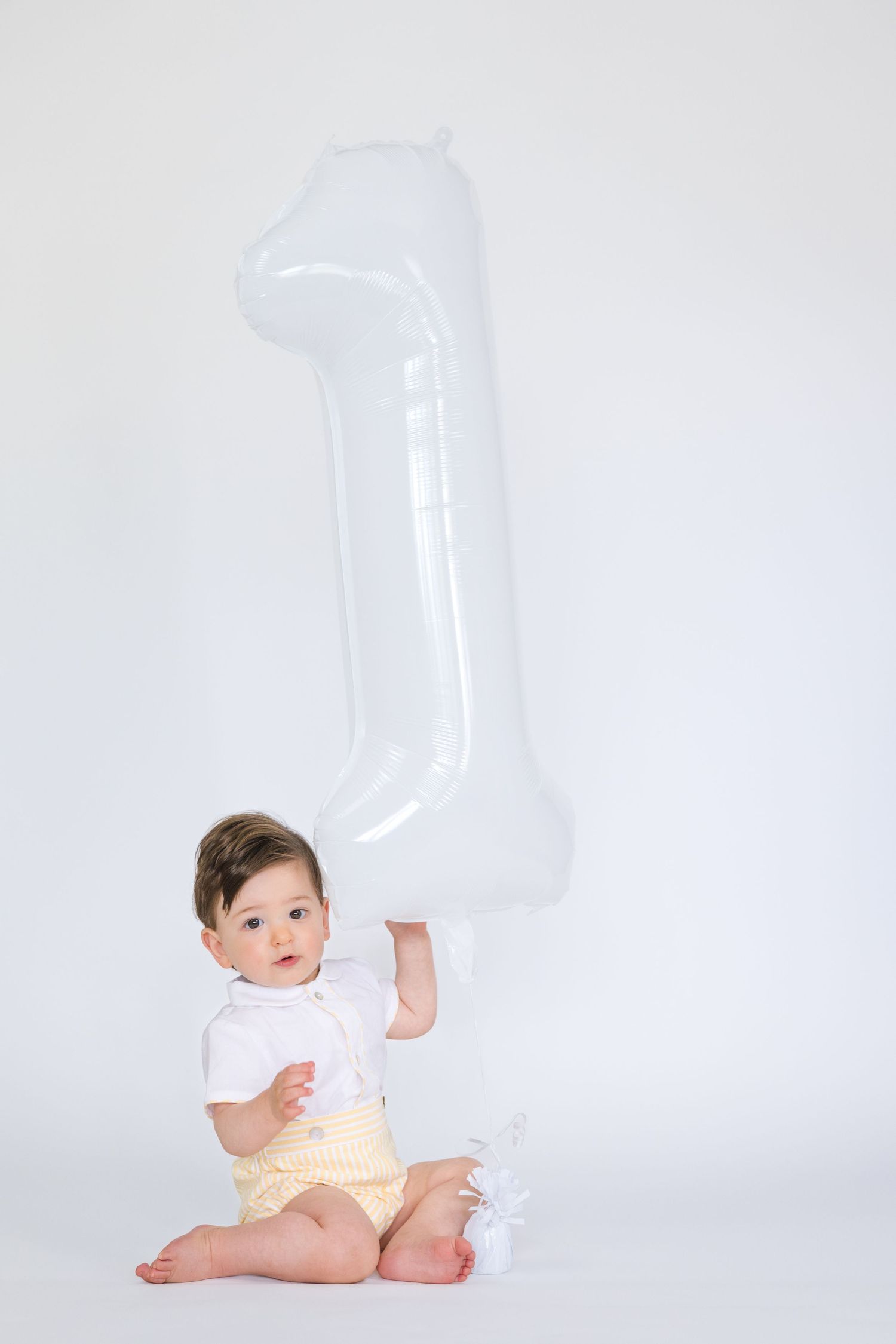 Baby sitting beside large transparent number one balloon against white backdrop.