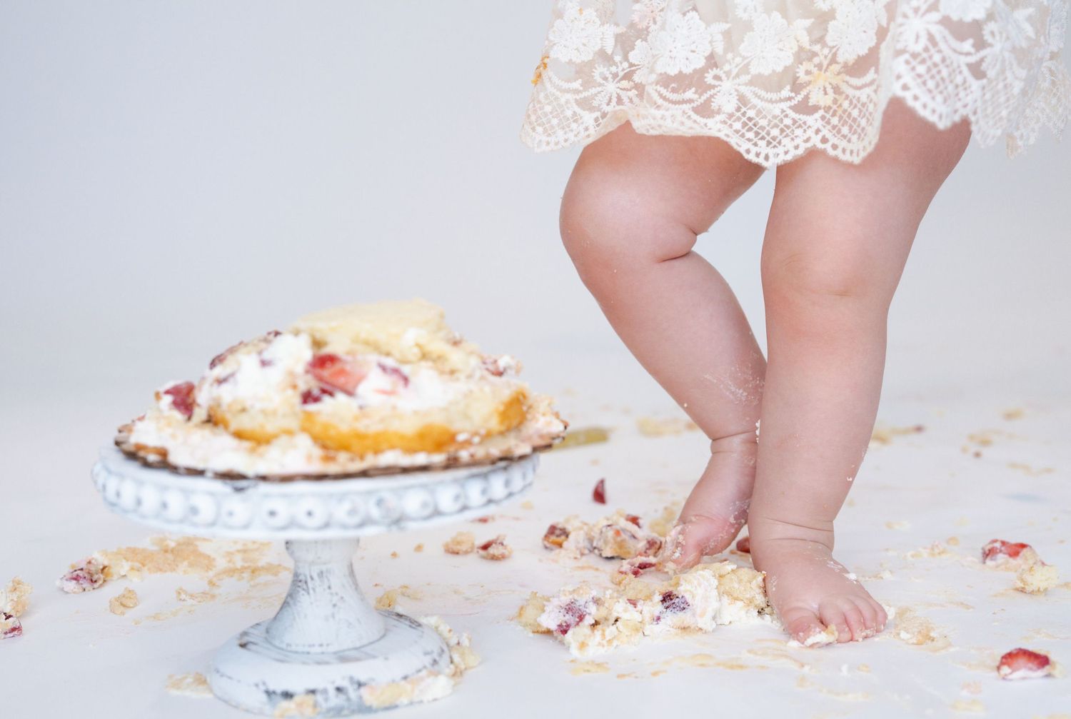 Small bare feet stand next to a decorated cake on a white pedestal with crumbs scattered around.