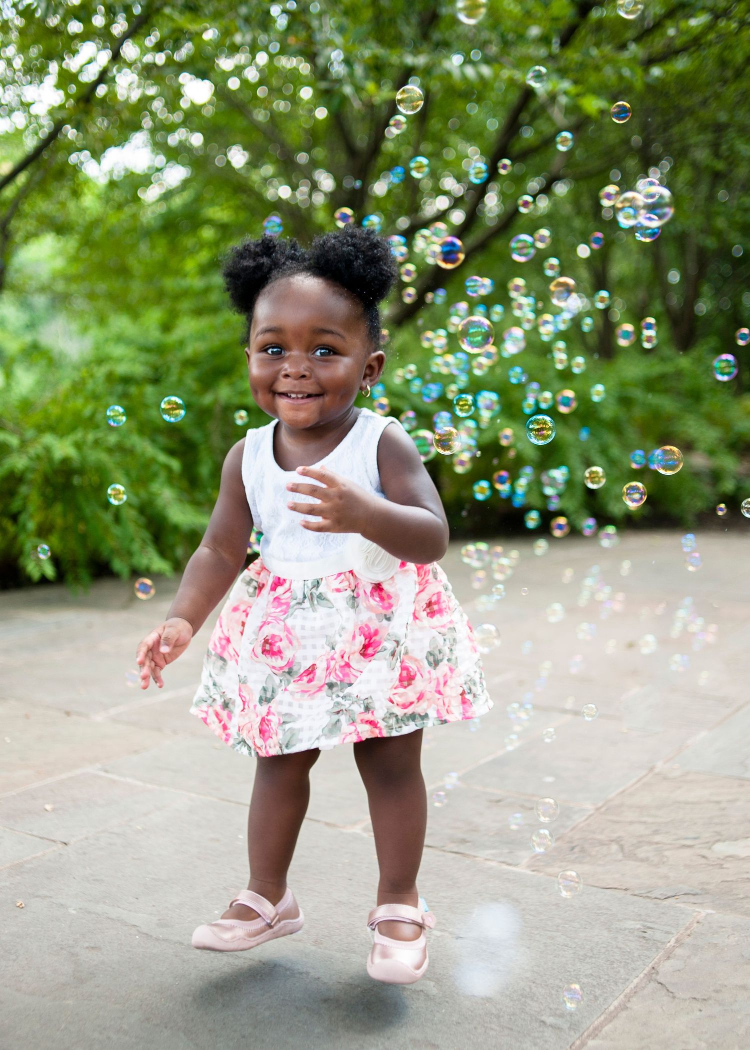 Child in floral dress dances among floating bubbles in garden setting.