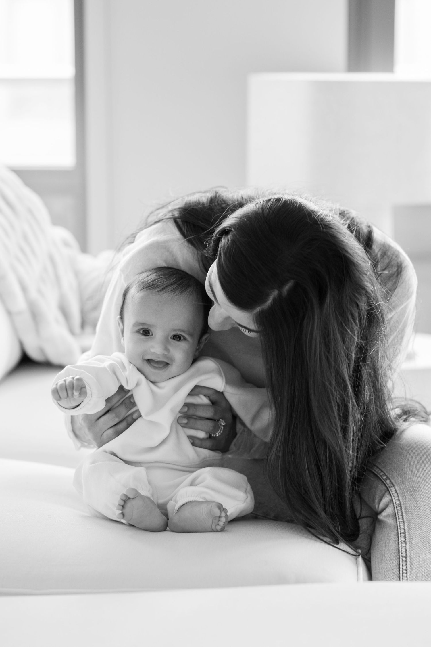 Black and white photo of loving mother kissing smiling baby on white sofa in bright room.