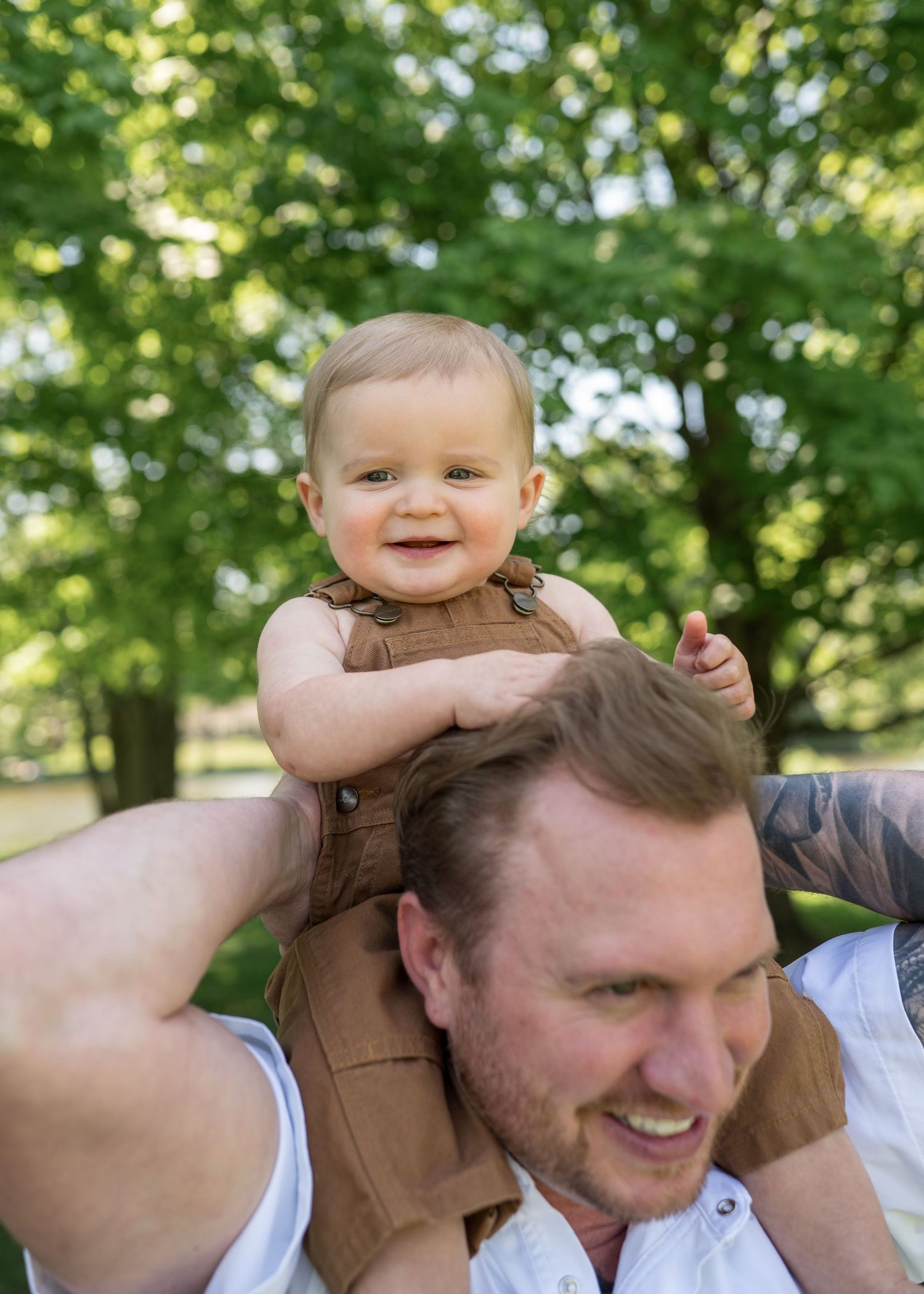 Happy baby in brown overalls sitting on fathers shoulders smiling in a green park.