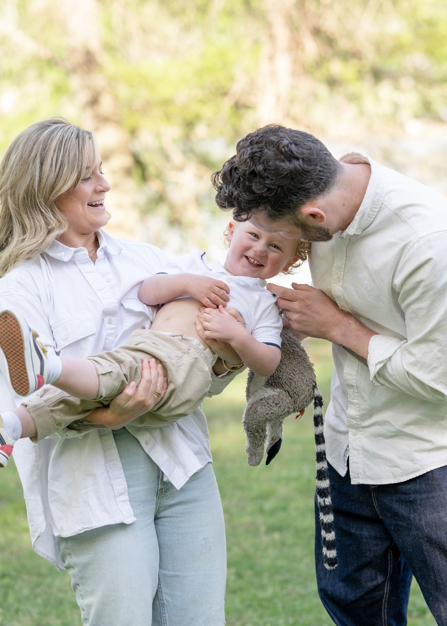 Happy family outdoors, mother holding laughing toddler while father playfully nuzzles child in sunny green park.