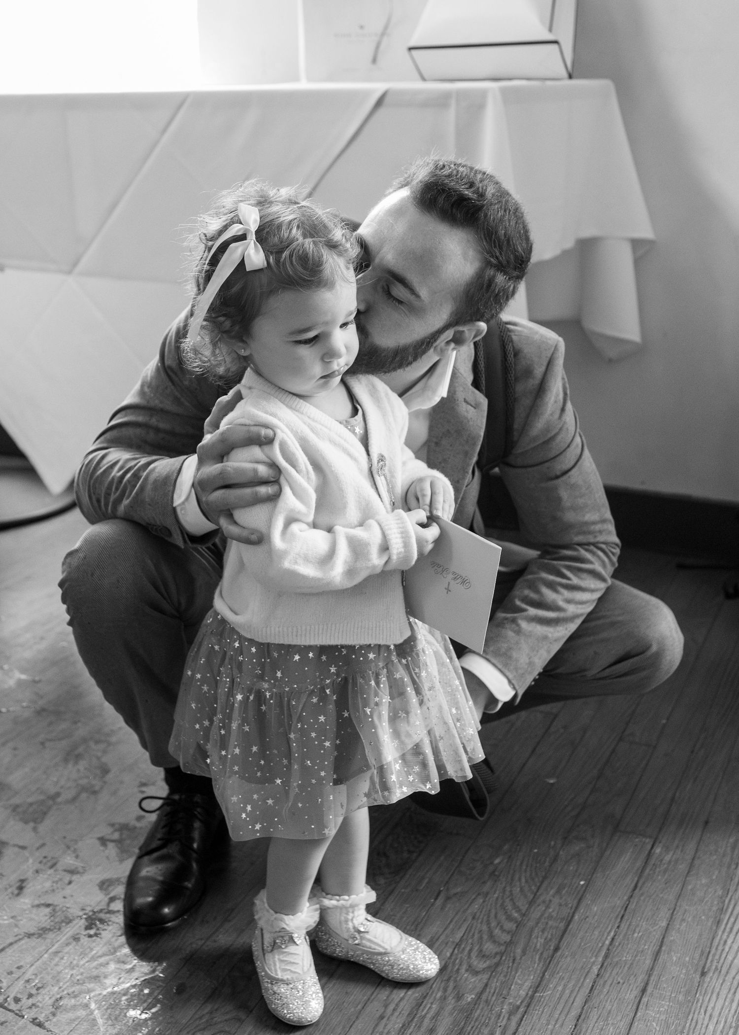Black and white photo of a bearded man crouching down to kiss a toddler girl in a tutu skirt and bow.