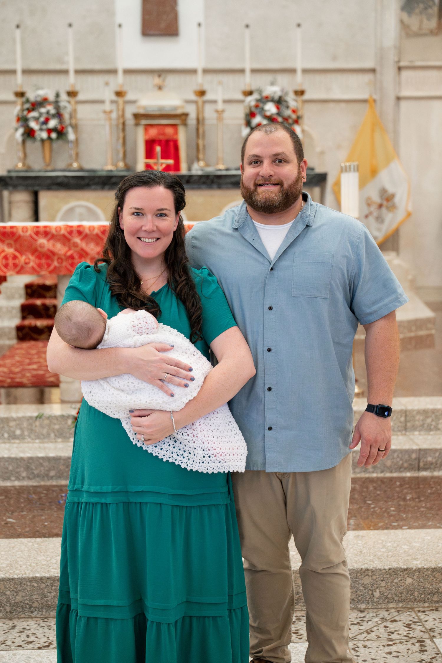 A couple in emerald green and khaki poses together with a baby wrapped in white lace in an outdoor setting.
