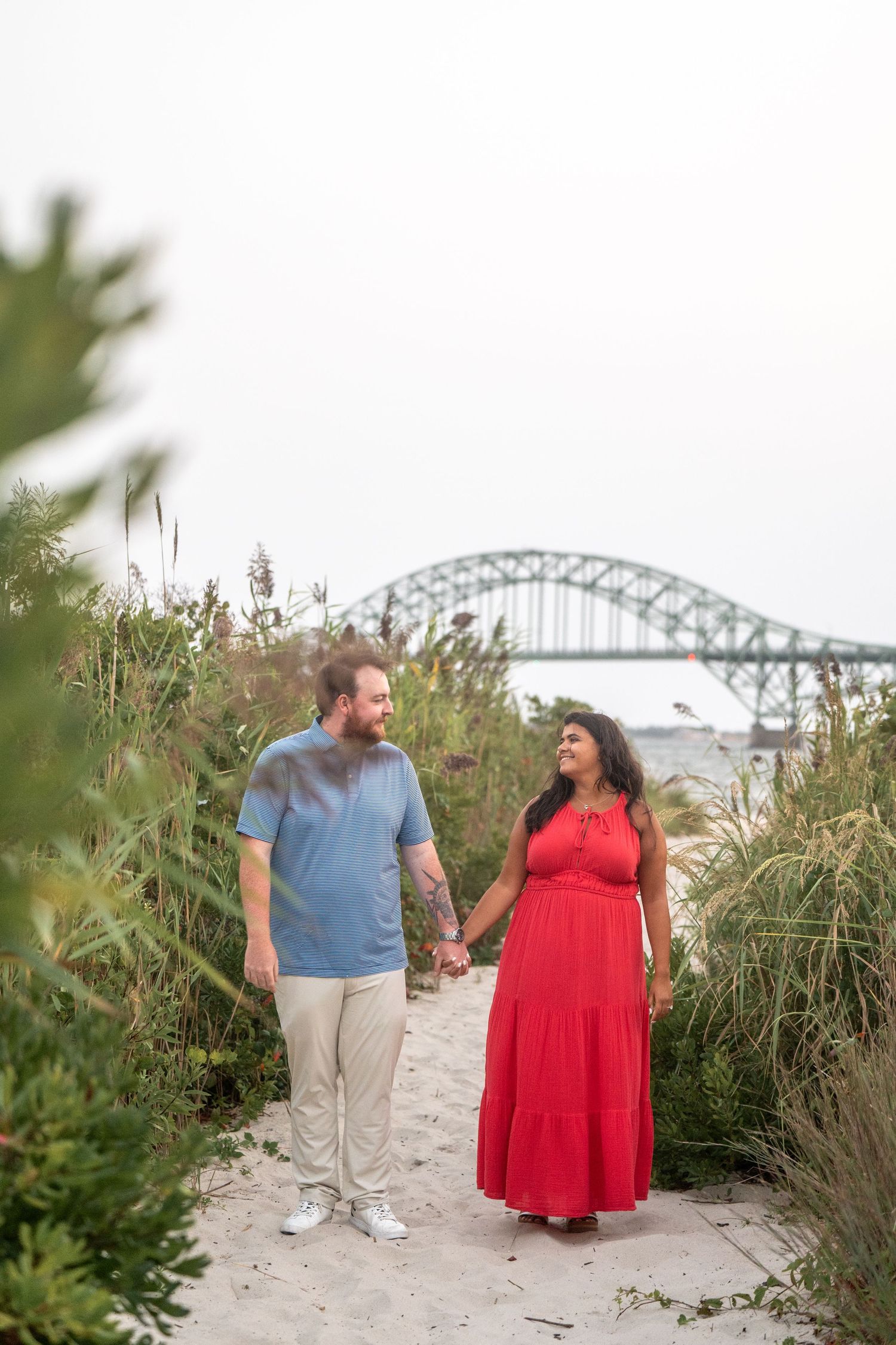 A couple shares a romantic moment on a beach path with a bridge visible in the background.