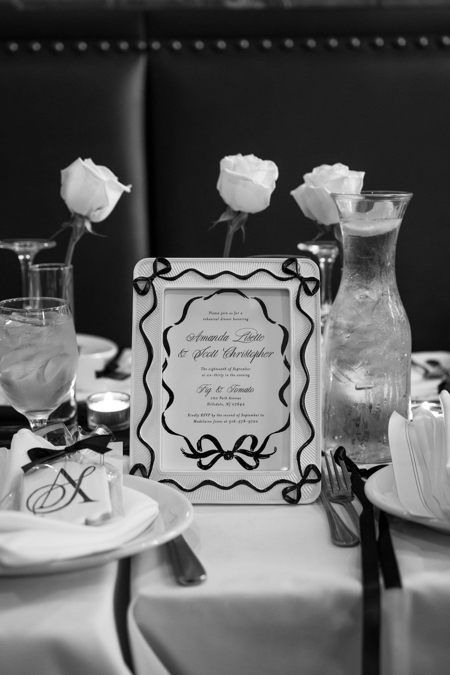 Black and white panoramic view of elegant table settings with framed menu cards and white roses at a formal event.