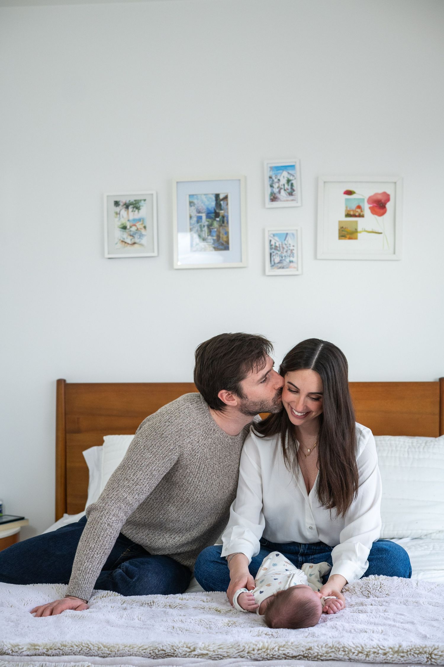 A tender family moment captured on a white bed with framed artwork on the wall.