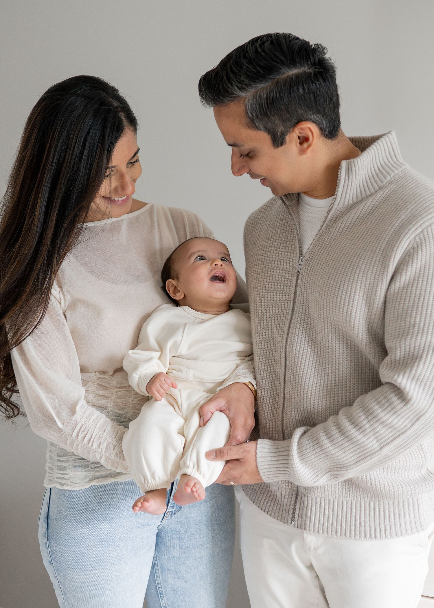 A couple in matching neutral sweaters and jeans share tender moments while holding their baby in a lifestyle photoshoot.