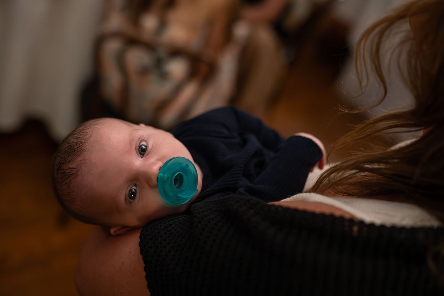 A baby lies on a white blanket while holding a turquoise pacifier in their mouth.
