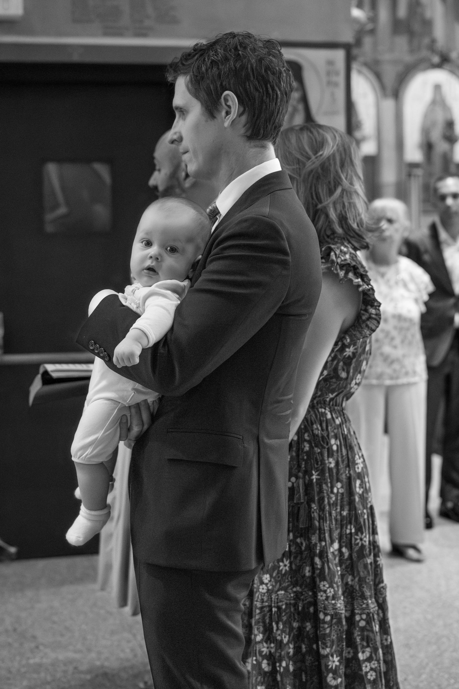 A baby in white clothing is held during an outdoor event as guests gather in formal attire.