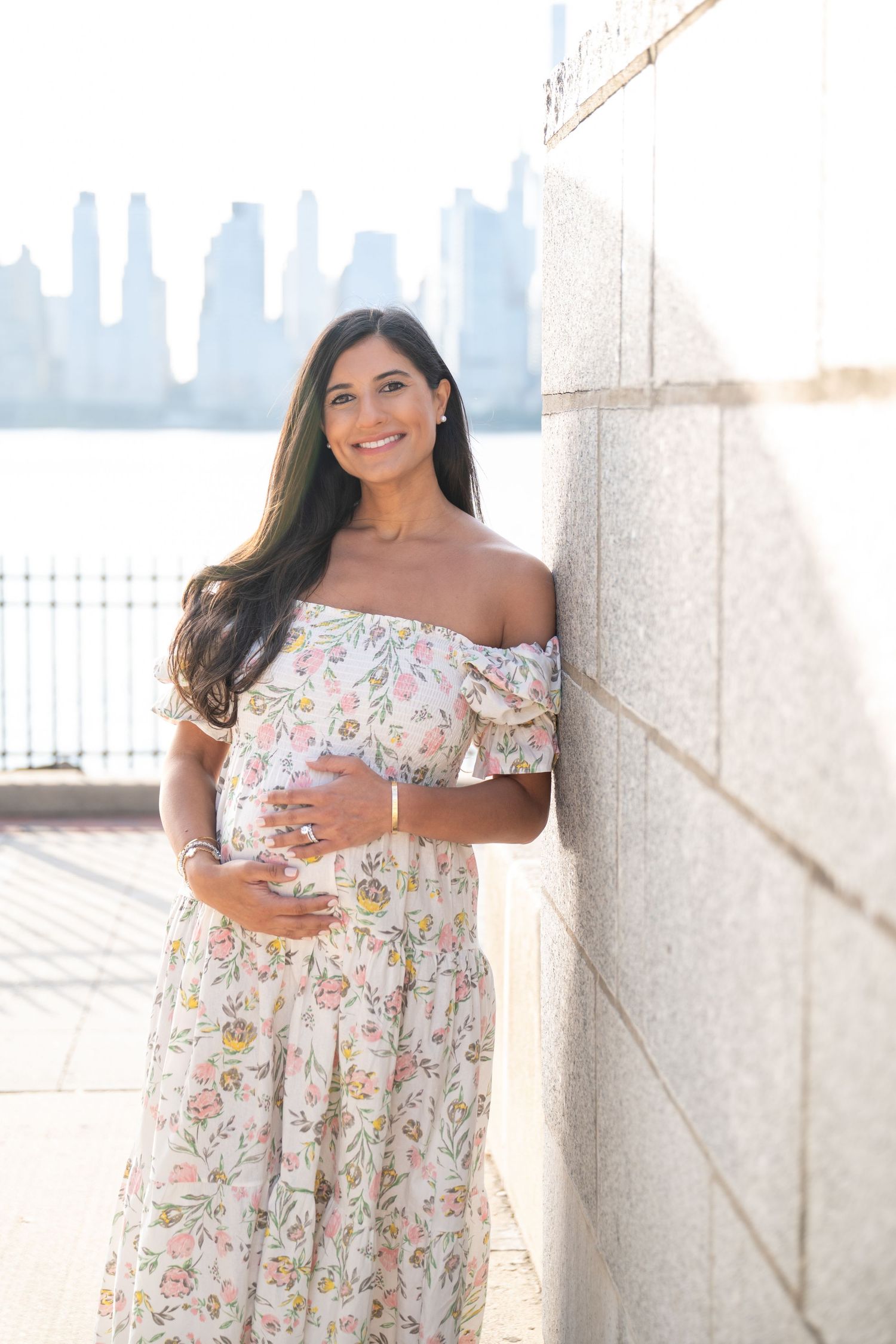Woman in a floral off-shoulder dress poses against a white wall with city skyline in background.