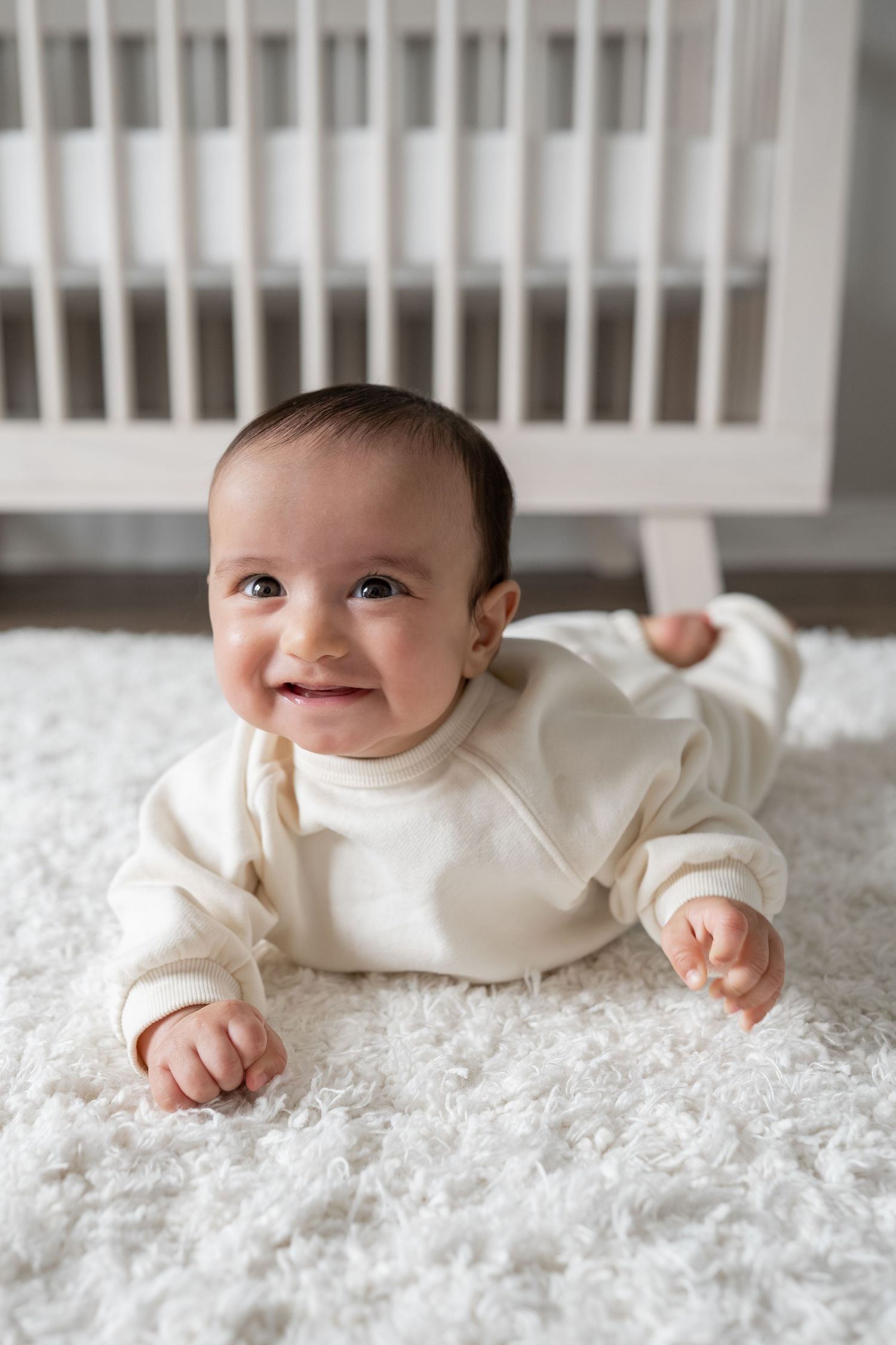 Smiling baby in cream outfit doing tummy time on white fluffy rug in front of white crib.