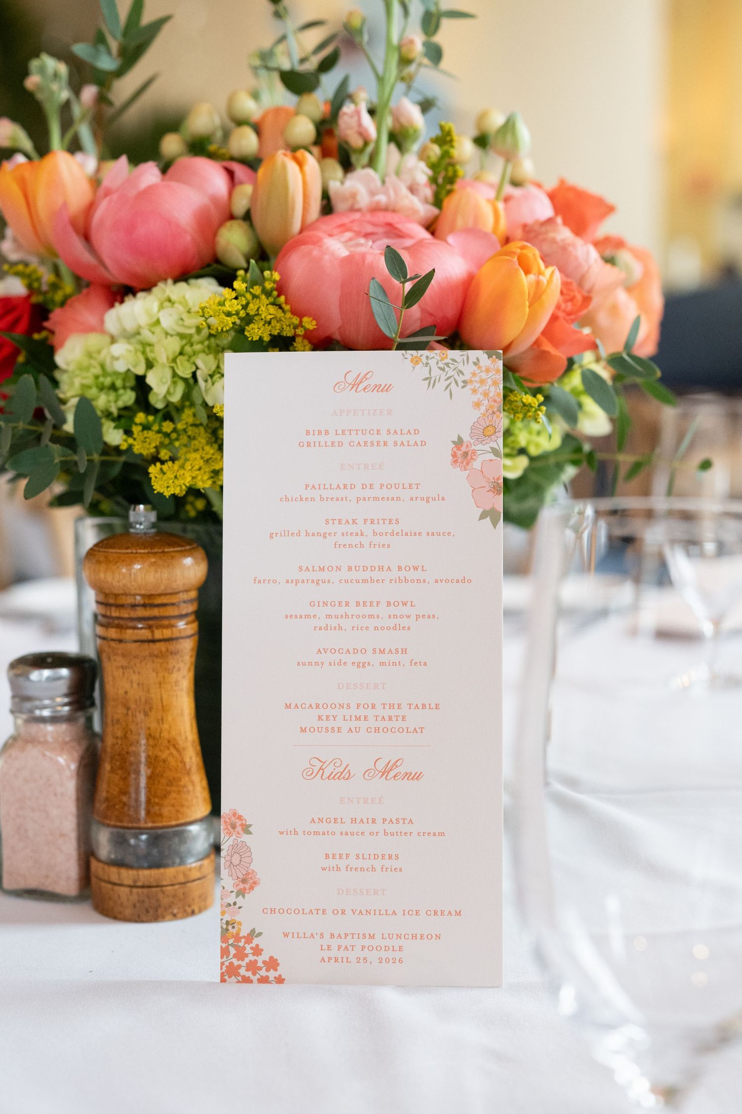 Elegant wedding menu card displayed beside a wooden pepper mill and vibrant floral centerpiece with pink peonies and orange tulips.