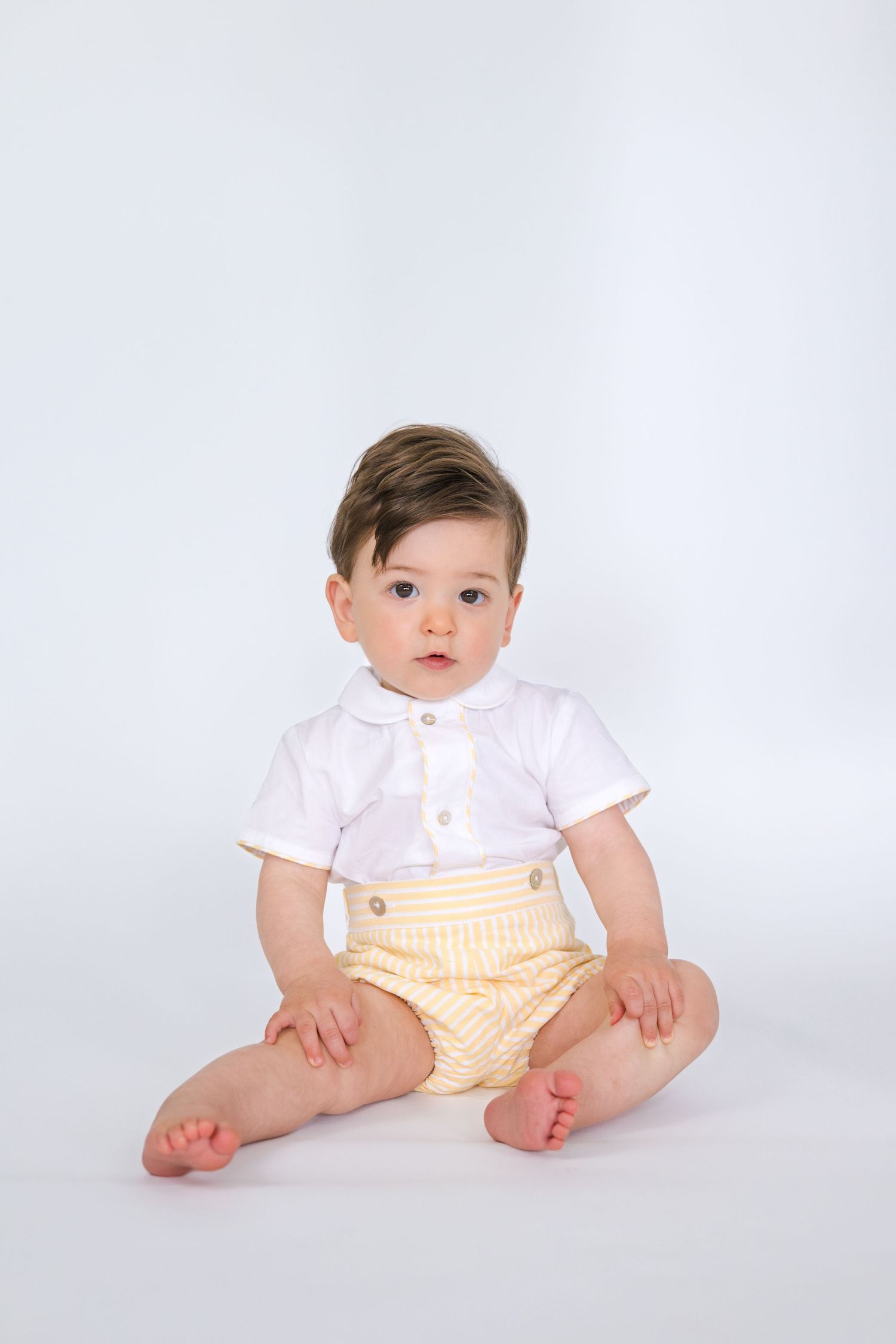 Baby wearing white collared shirt and yellow striped bottoms sitting against white background.