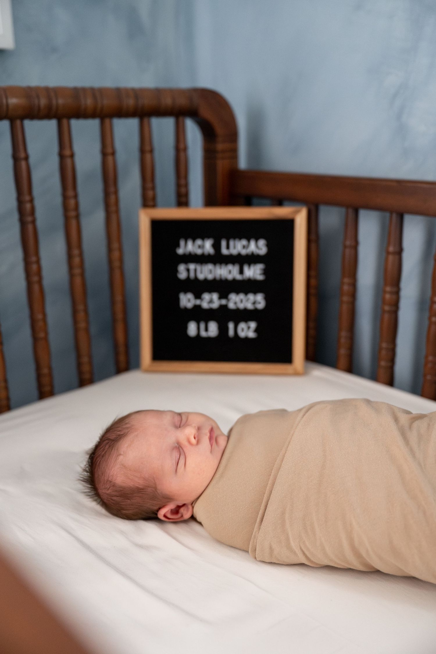 A sleeping newborn baby in a crib with a black letterboard displaying birth details.