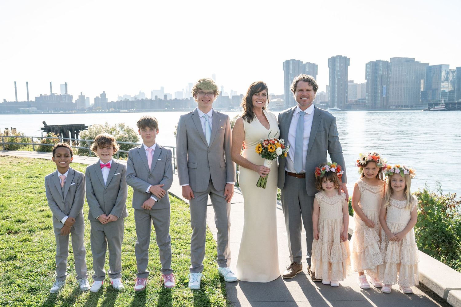 Wedding party in light gray suits and blush dresses poses together on a sunny waterfront with city skyline in background.