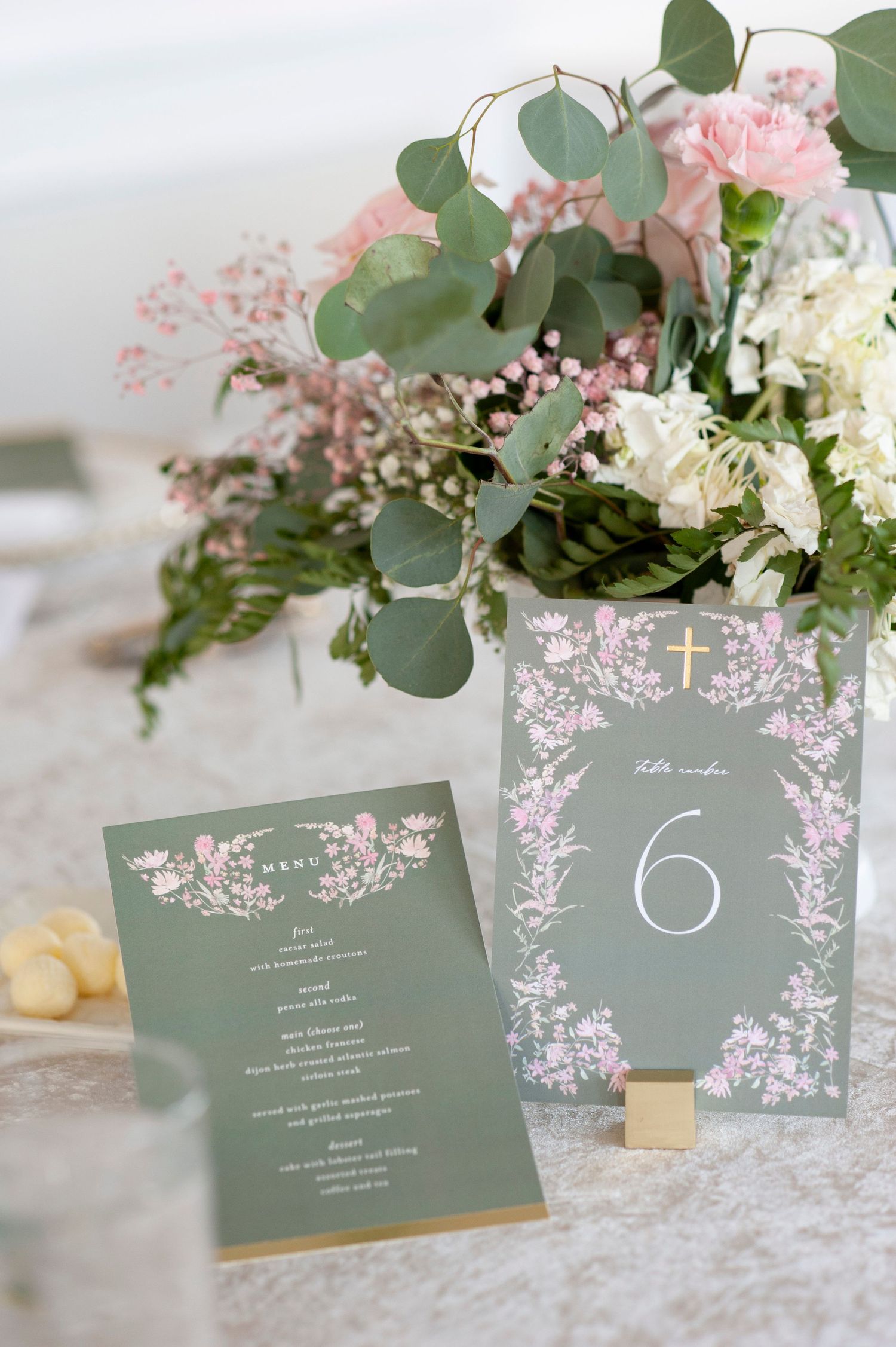 Elegant sage green wedding stationery with floral designs displayed alongside eucalyptus and pink flower arrangements.
