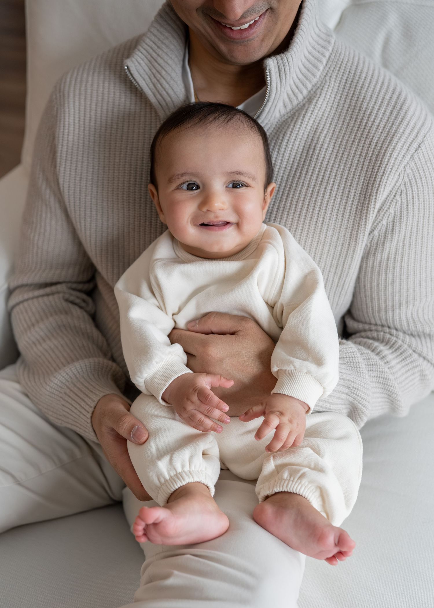 Smiling baby in cream outfit sitting on mother's lap, both wearing neutral tones in a bright, cozy setting.