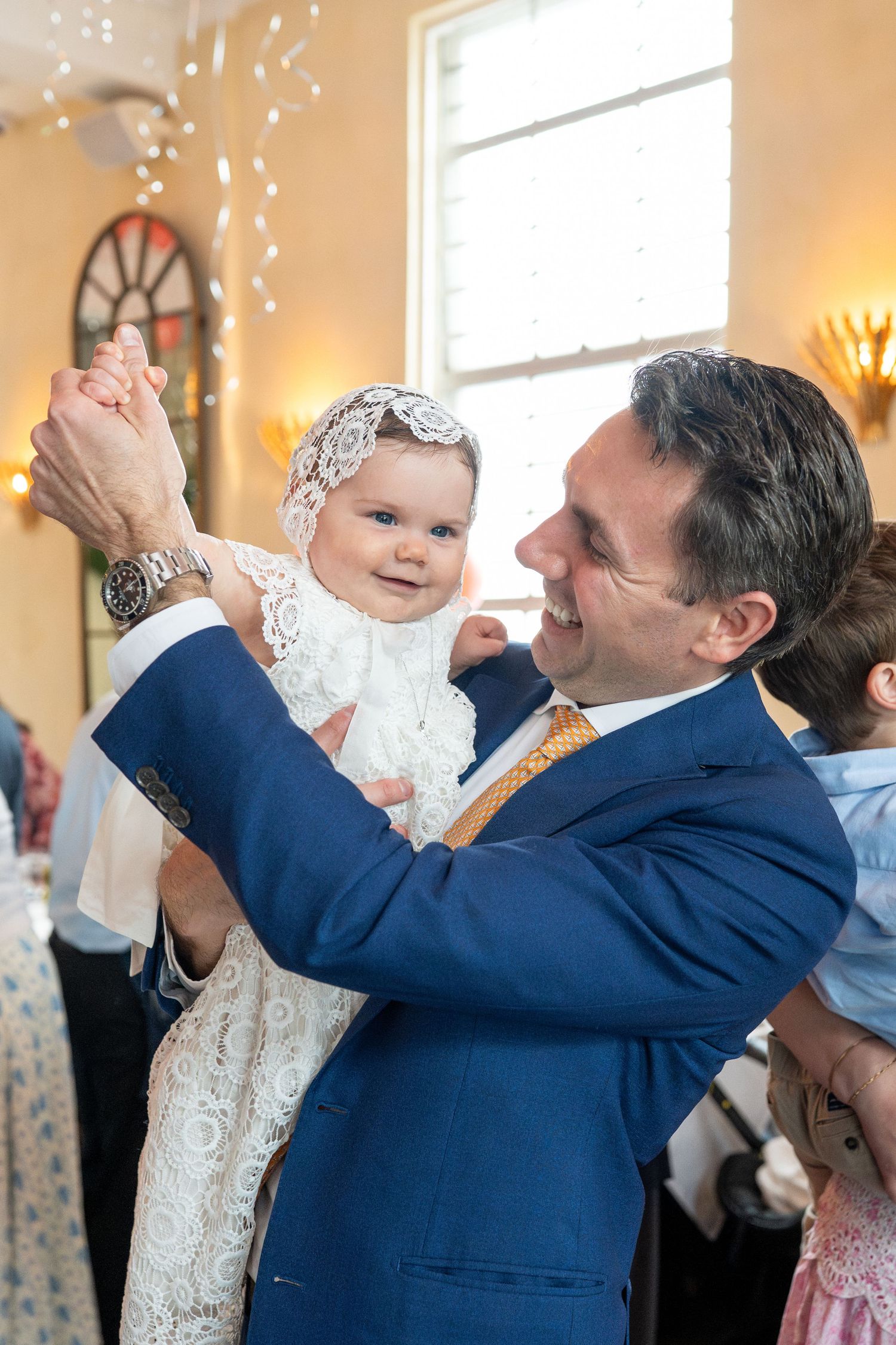 Father in blue suit holding baby in christening gown during baptism celebration.