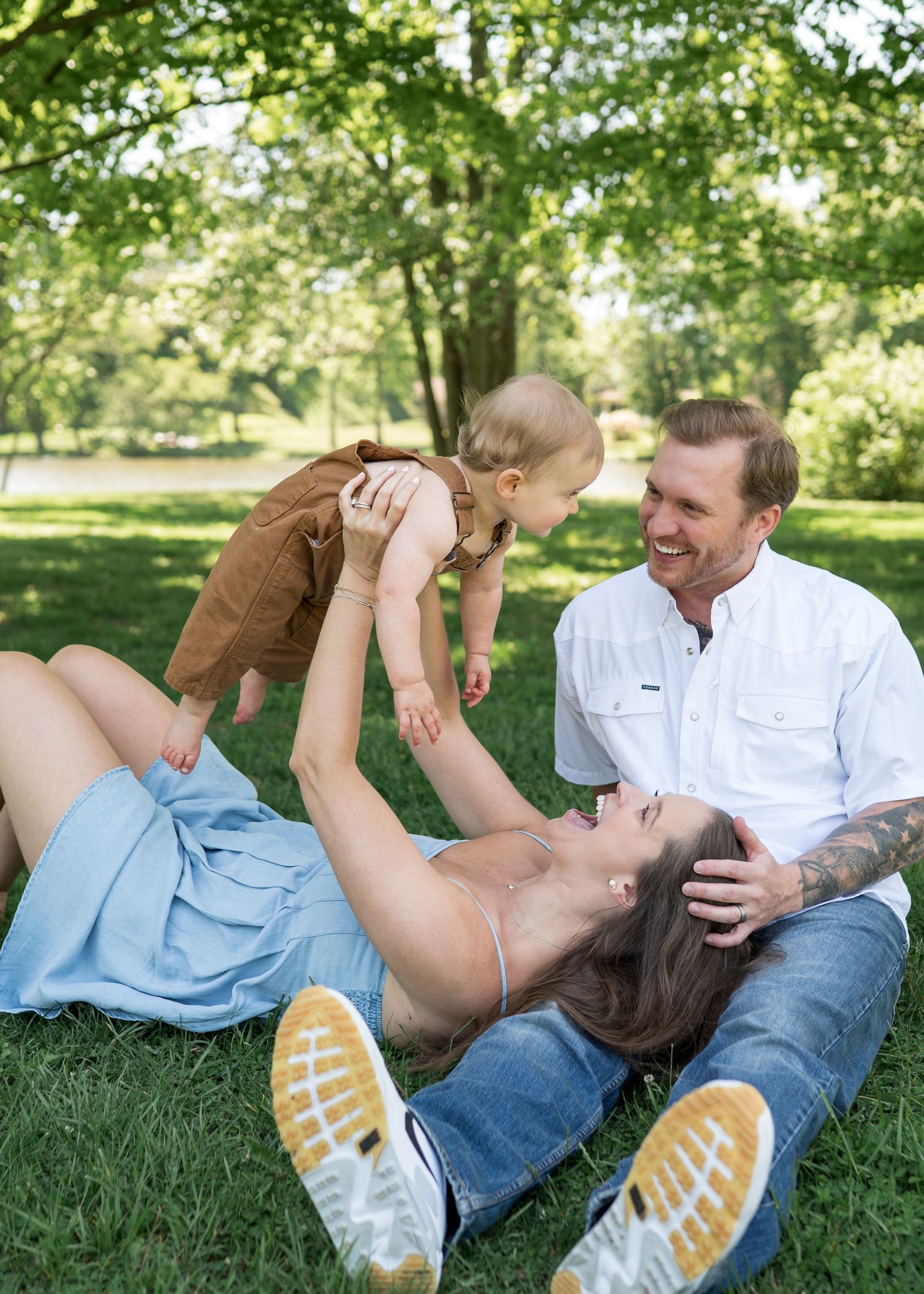 A happy family enjoys a sunny day in the park, with mom lifting baby while dad smiles beside them on the grass.