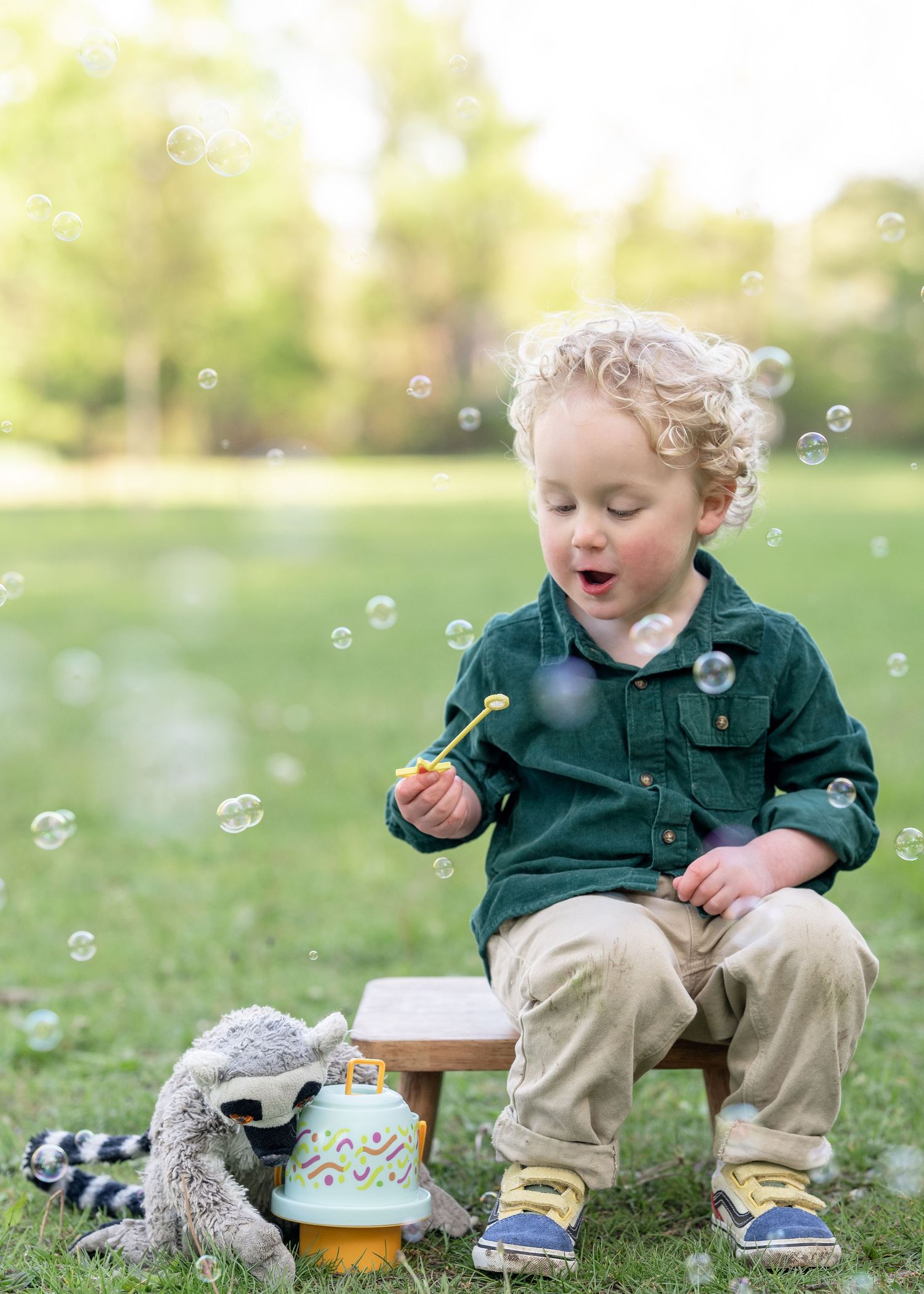 Curly-haired toddler boy blowing bubbles outdoors on a wooden stool with stuffed animals nearby.