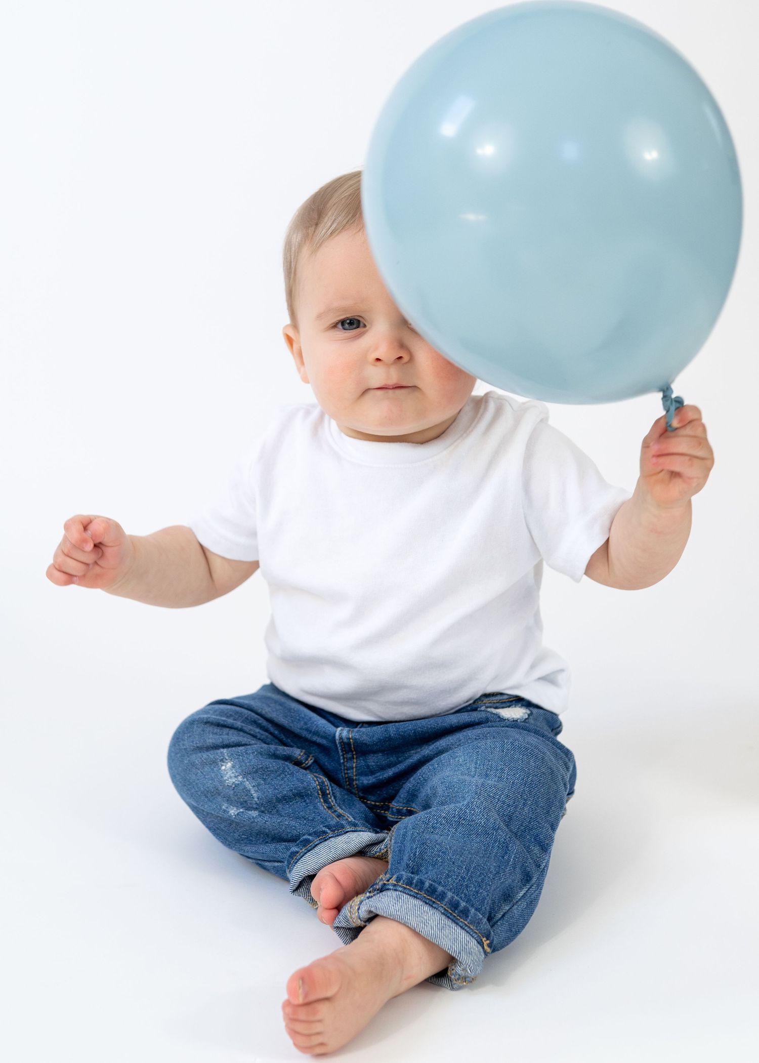 Cute baby in white onesie and jeans sitting on white background holding a light blue balloon.