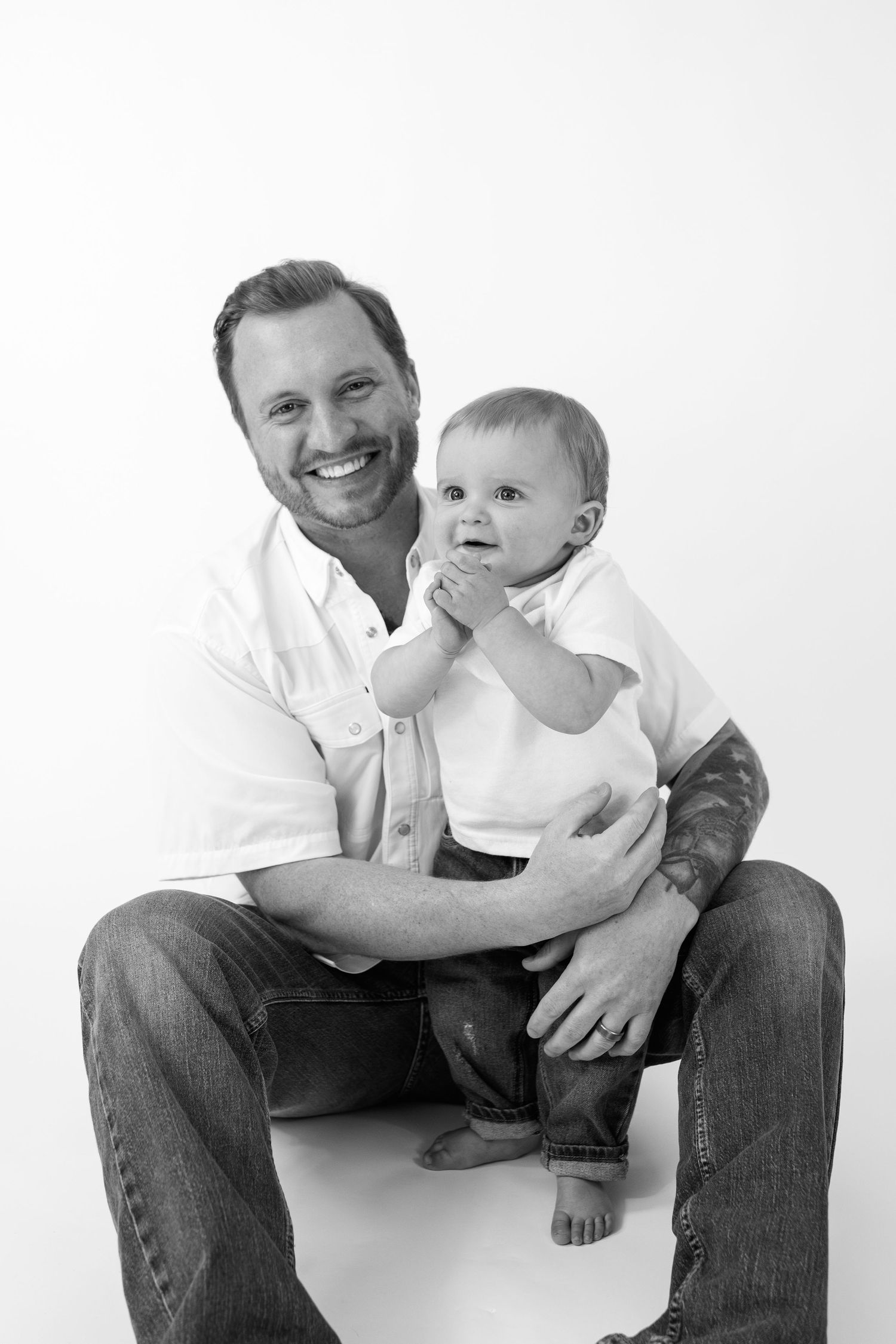Black and white portrait of smiling father holding baby in white onesie, both wearing jeans on white background.