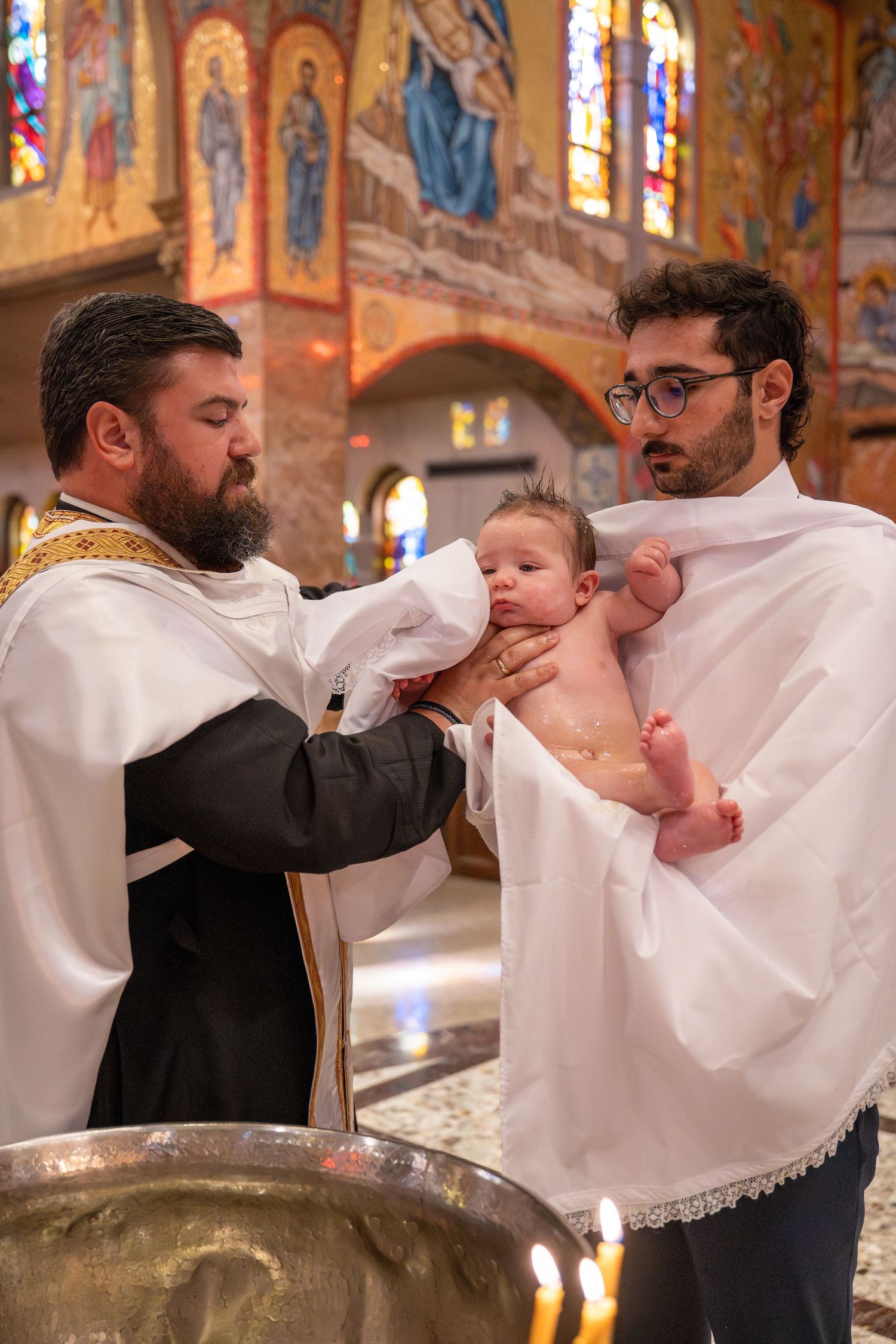 A religious baptism ritual being performed at a church altar with colorful stained glass windows in the background.