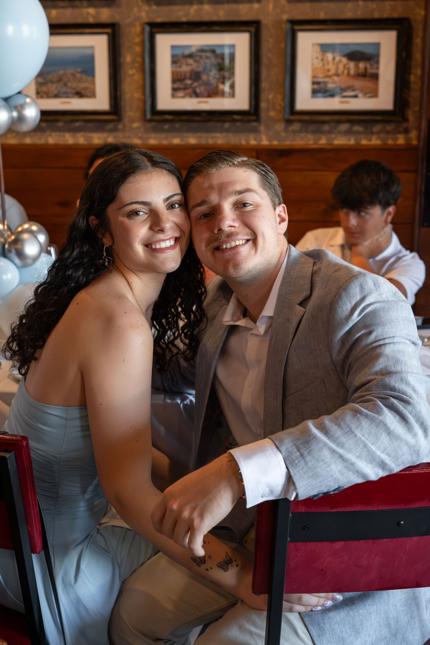 A couple in formal wear share a joyful embrace while seated in wooden chairs at an event.