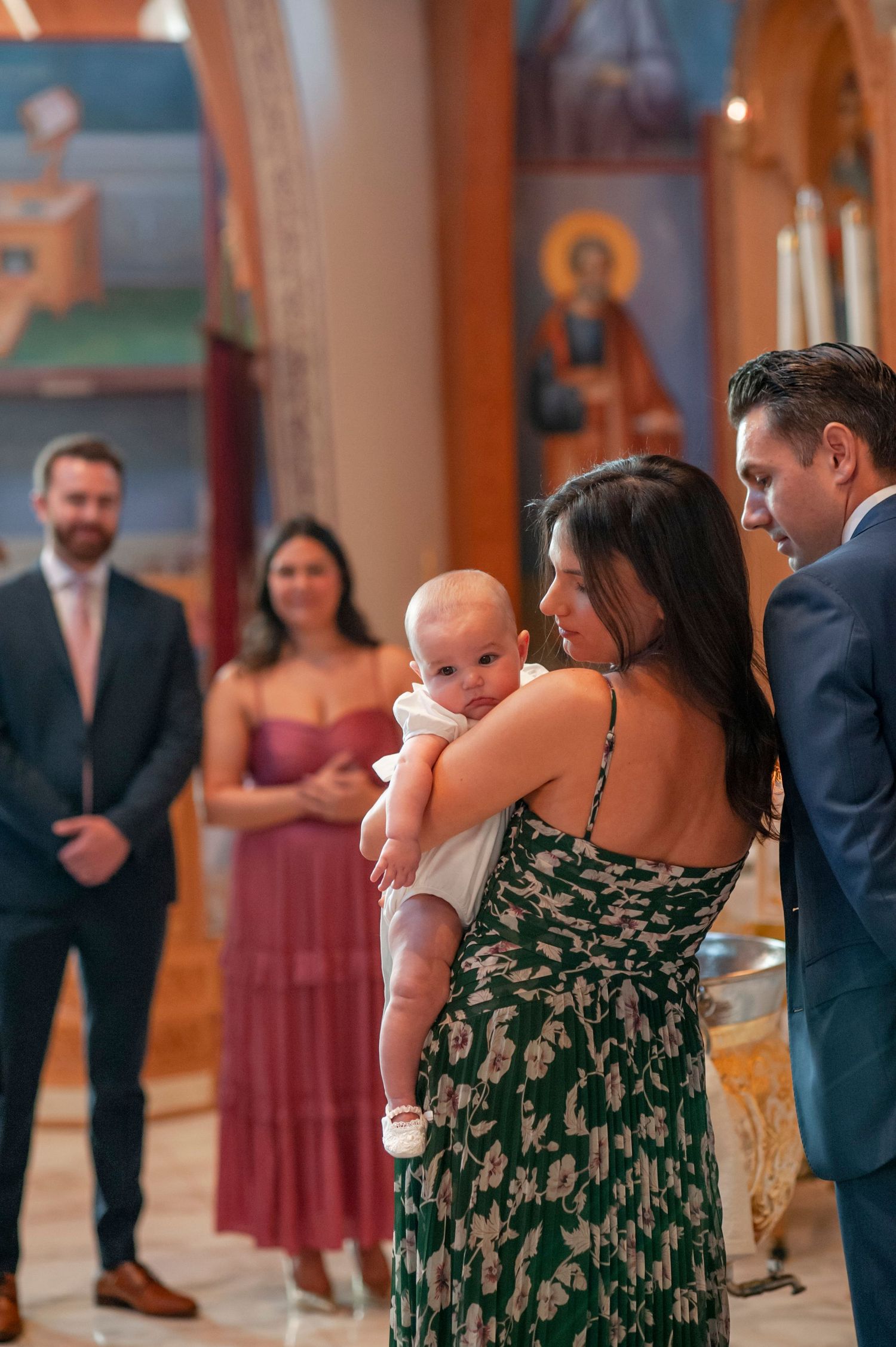 A baptism ceremony in a church with attendees gathered around a baby wearing white, surrounded by religious icons.