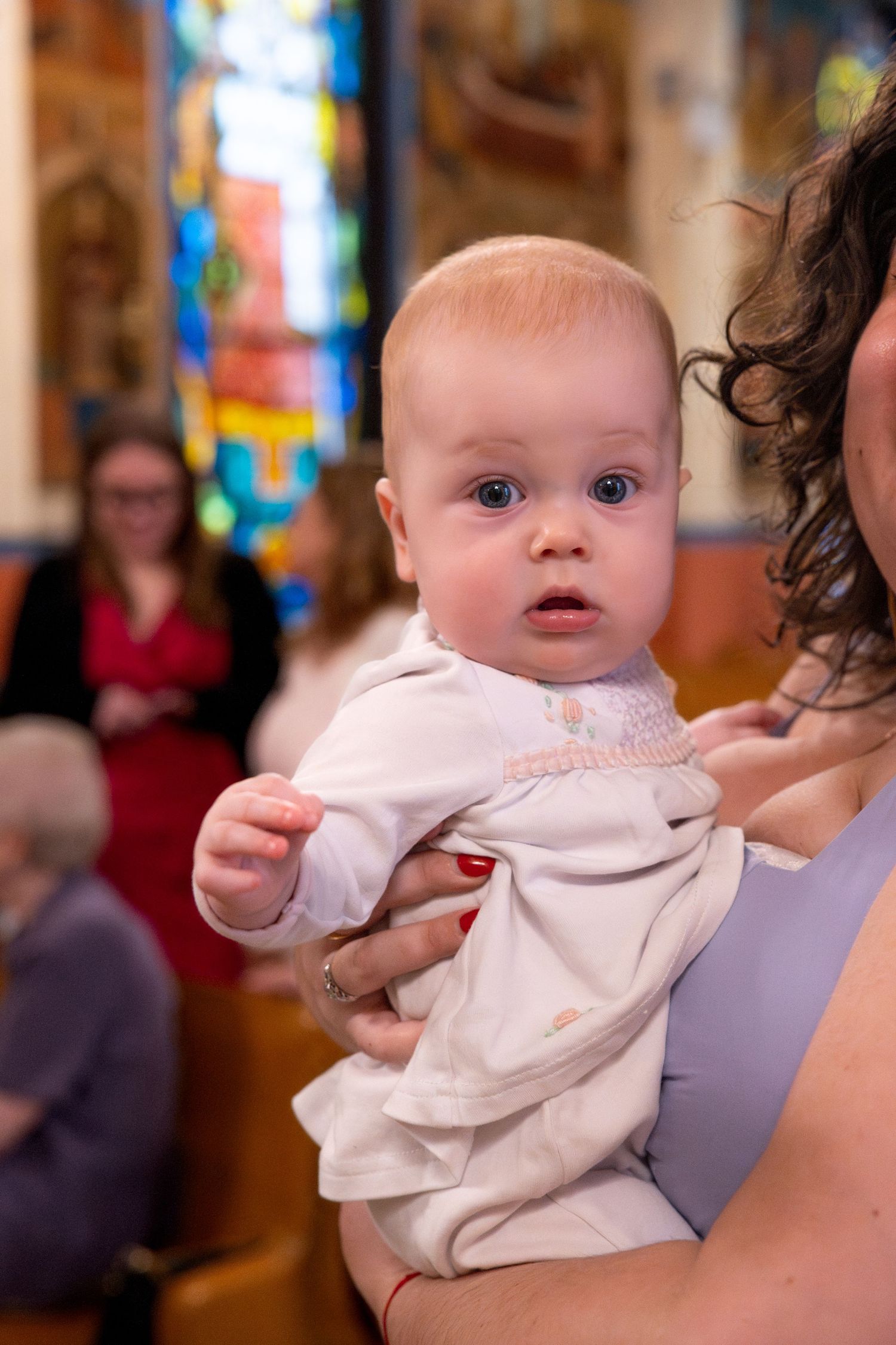 A baby in a white christening gown is held during a baptism ceremony in a church with stained glass windows.