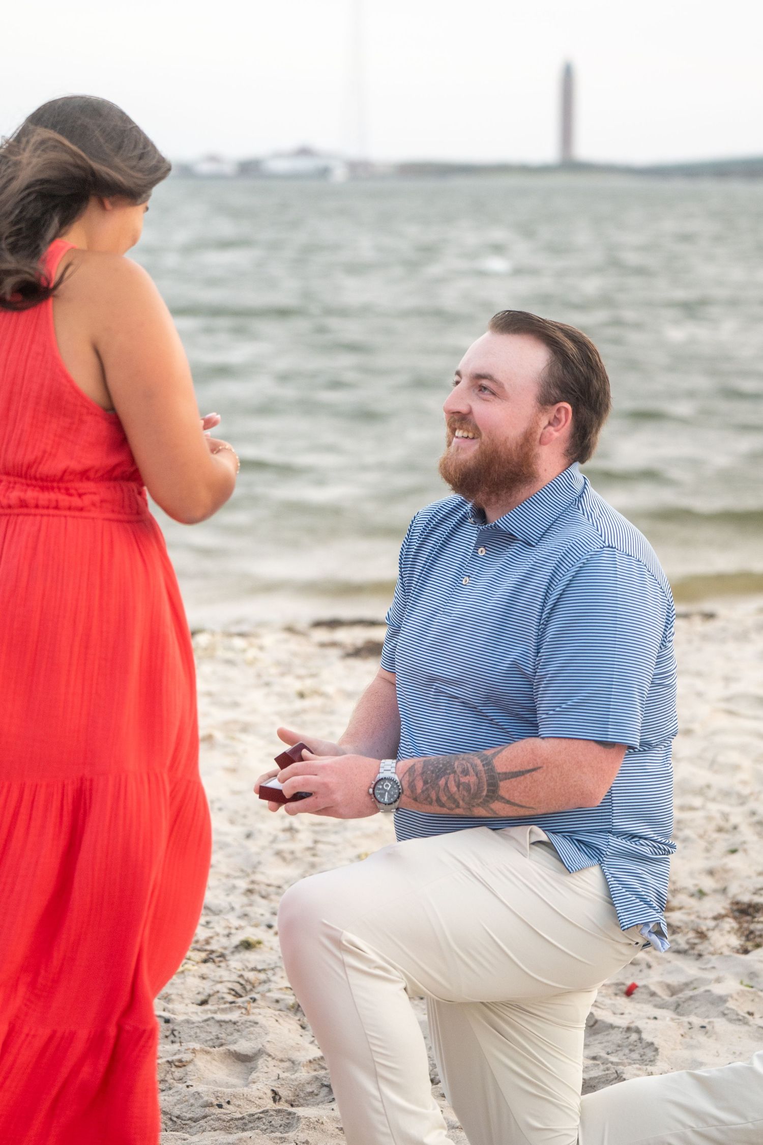 A couple shares a romantic beach proposal moment with the man kneeling in khakis and blue shirt by the ocean.