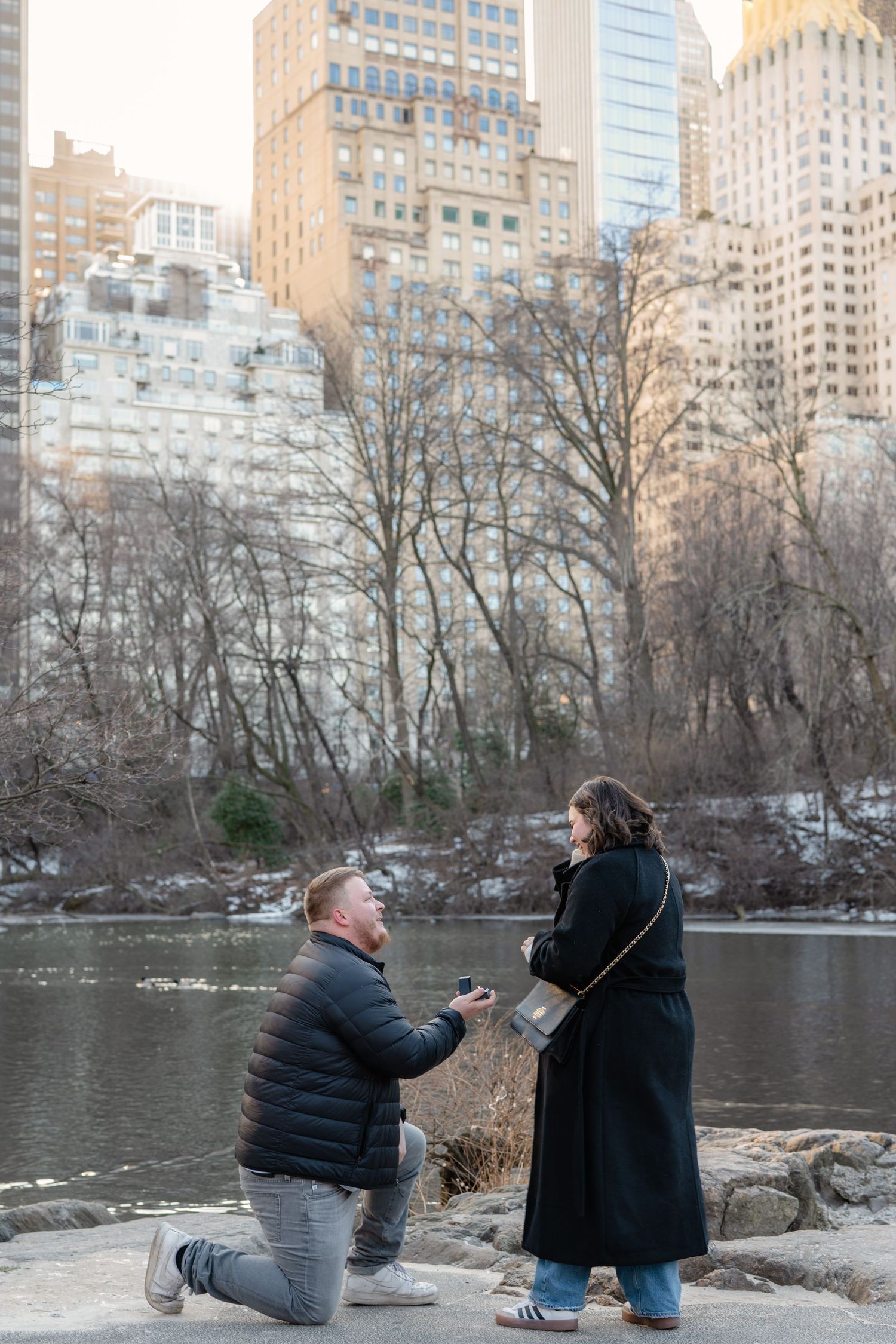 A couple shares a romantic proposal moment by a lake in Central Park with Manhattan buildings in the background.